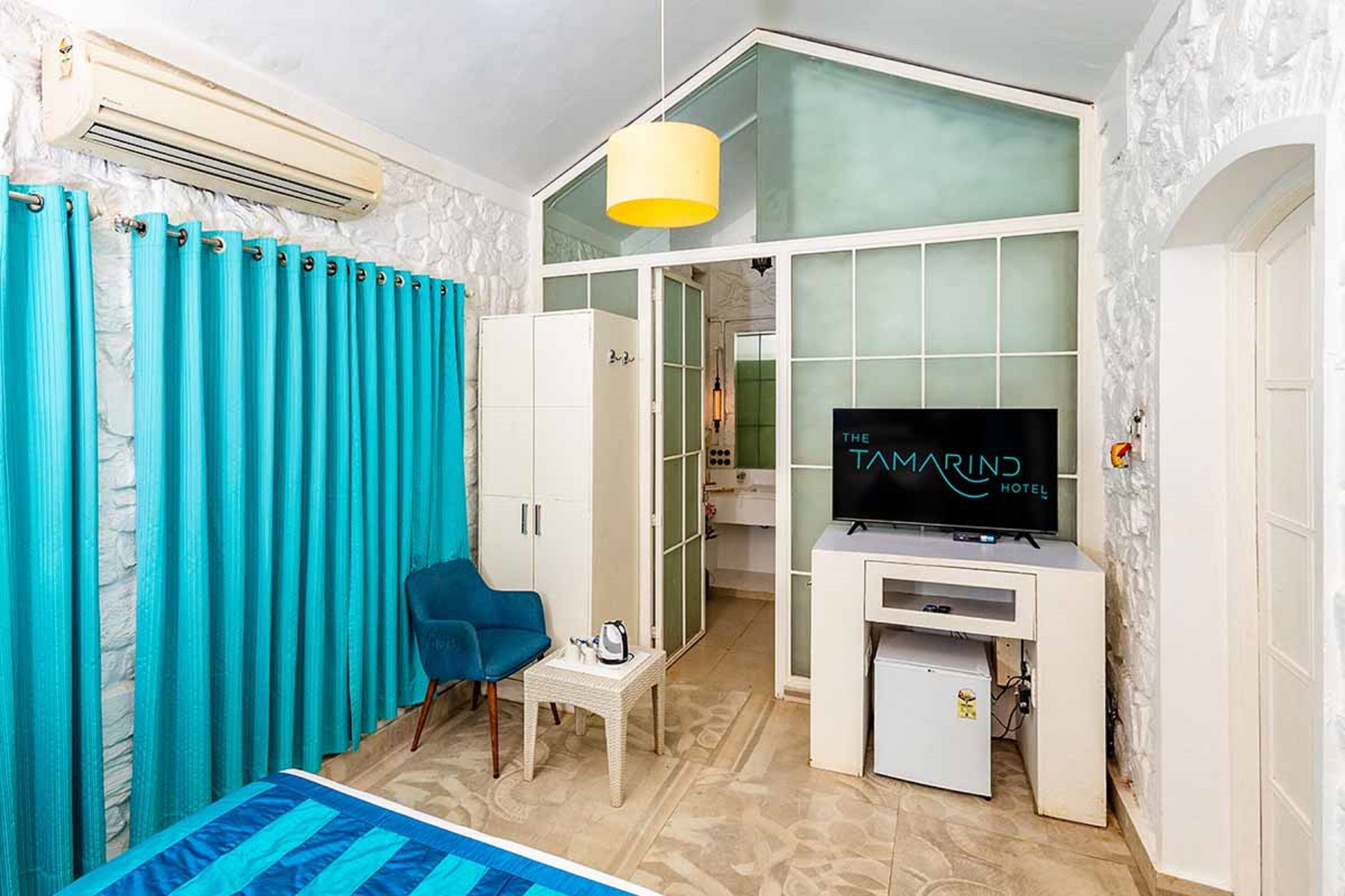 A hotel room with a bed, a TV, a small table, and a chair. There is a window with curtains and a door leading to a bathroom.