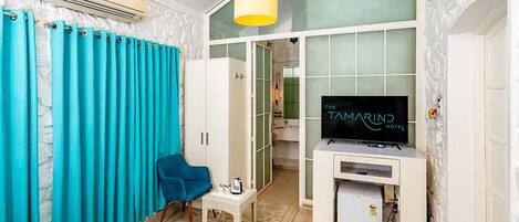 A hotel room with a bed, a TV, a small table, and a chair. There is a window with curtains and a door leading to a bathroom.