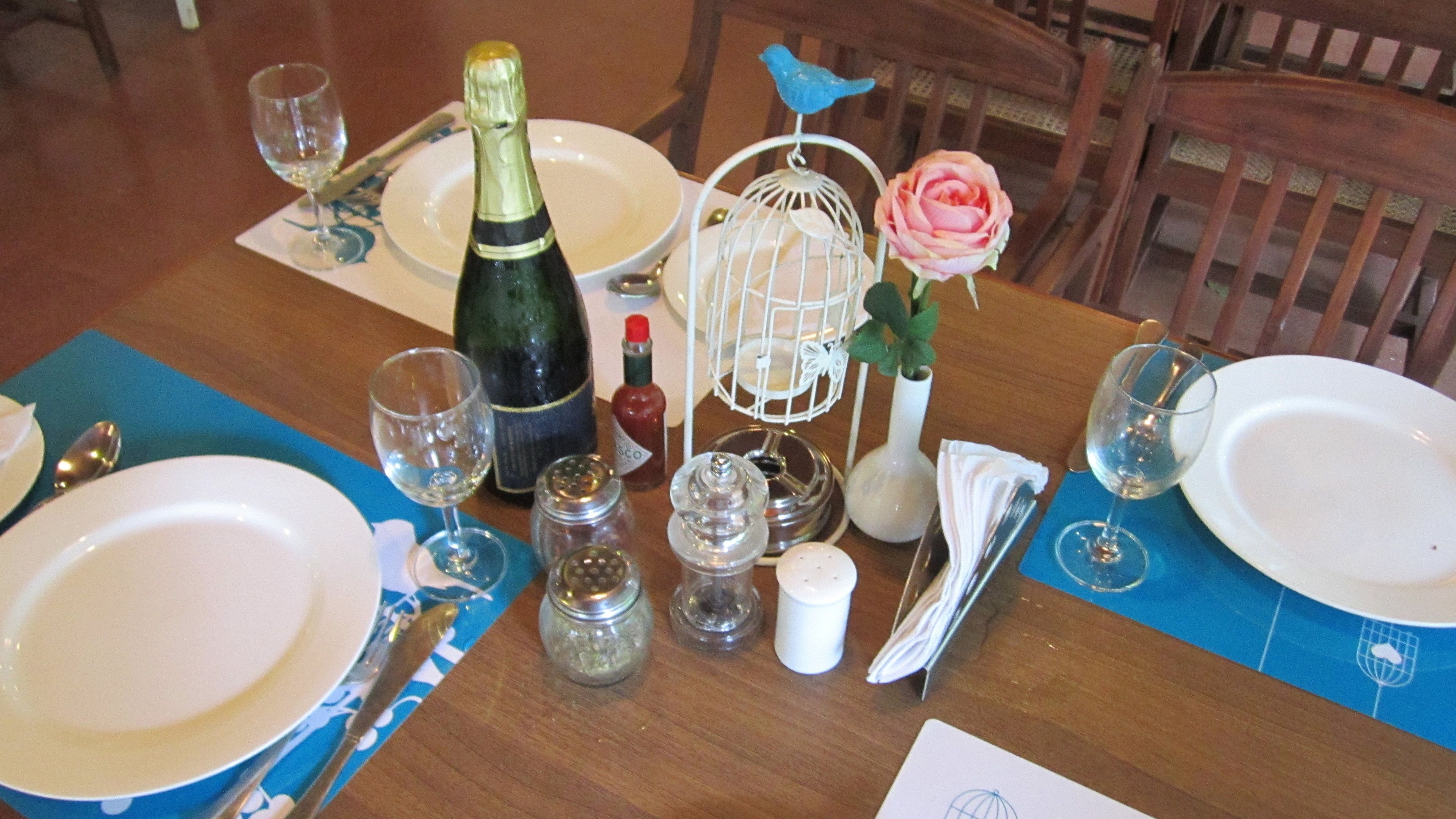 Una mesa de comedor dispuesta con platos, cubiertos, vasos, una botella de champán, saleros y pimenteros, una jaula con una rosa y una servilleta.