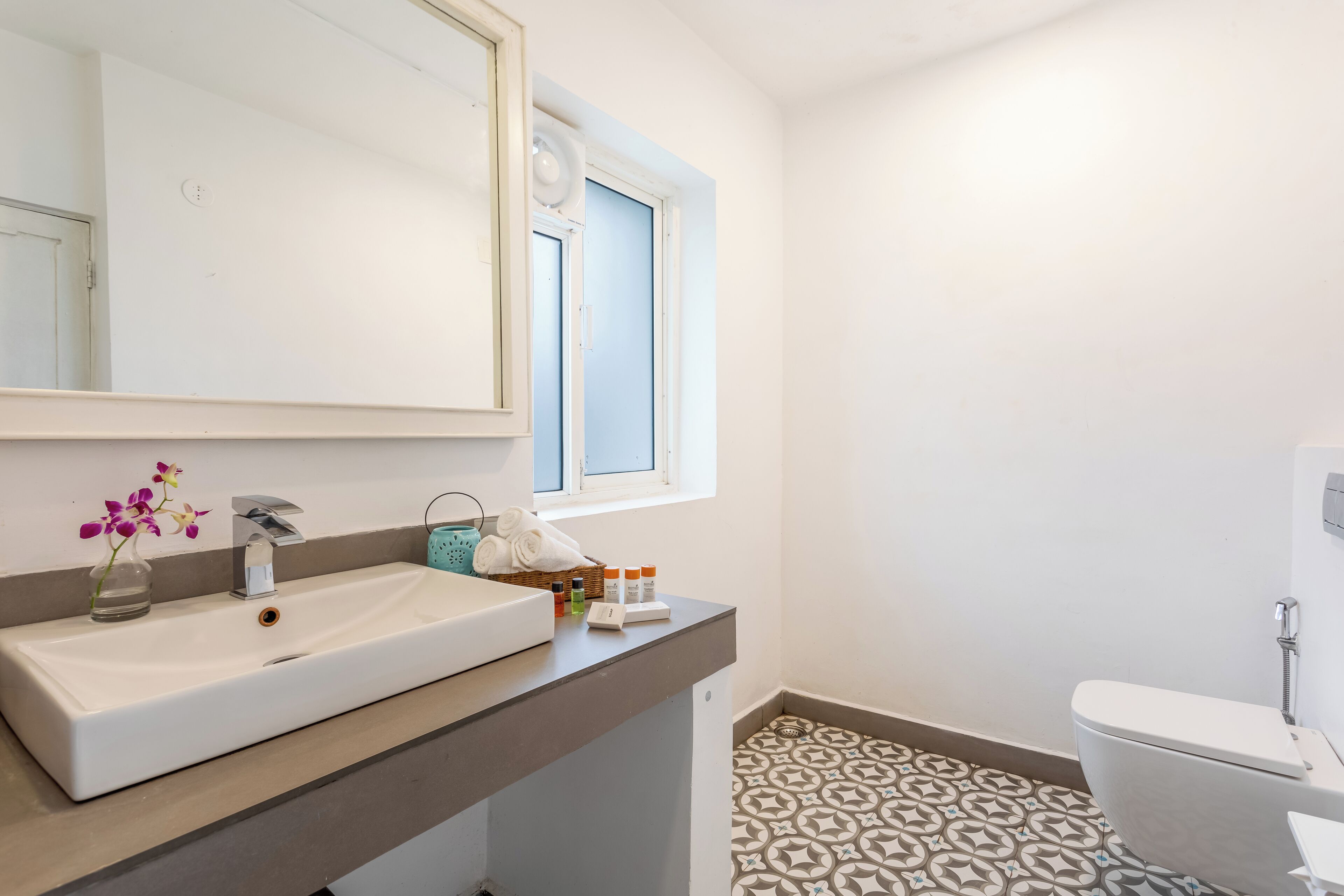 Un baño con lavamanos blanco, espejo, ventana, inodoro y baldosas de suelo estampadas.