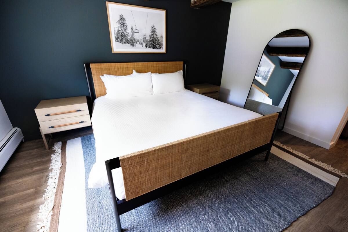 A neatly arranged bedroom with a large bed, a bedside table, a nightstand, a mirror, and a framed picture on the wall.