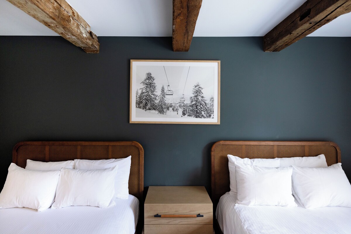 A room with two beds, a small wooden nightstand, and a framed picture of a snowy landscape.