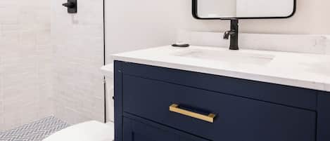 A modern bathroom with a white toilet, a dark blue vanity with gold handles, and a mirror with three lights.