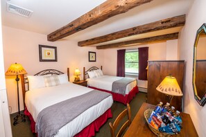 A hotel room with two beds, wooden beams on the ceiling, a window with curtains, and a table with a basket of toiletries. - The Lodge at Lincoln Peak (Warren)