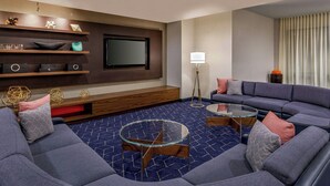 A modern living room with a sectional sofa, a glass coffee table, a flat-screen TV mounted on the wall, and a decorative shelf with various items. - Courtyard Oneonta (Oneonta)