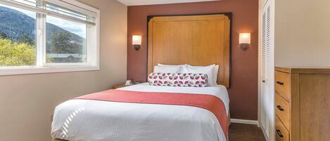 A hotel room with a large bed, a wooden headboard, two wall-mounted lights, a window with a view of mountains, and a wooden wardrobe.