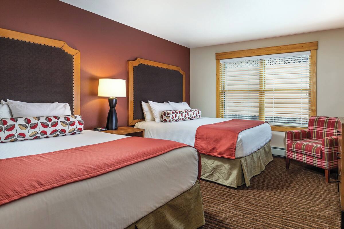 A hotel room with two beds, a desk, a chair, and a large window with blinds.