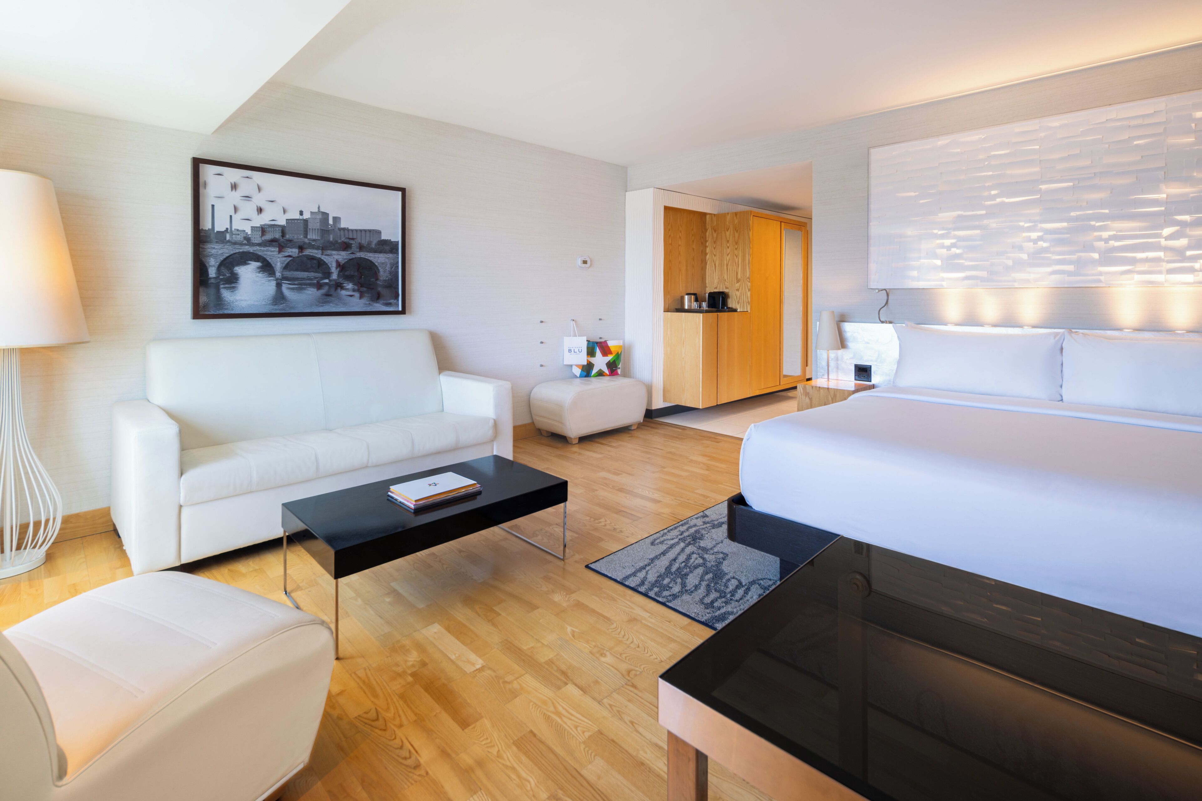 A modern hotel room with a bed, a sofa, a coffee table, and a desk. There is a cityscape painting on the wall, a wooden cabinet, and a lamp.