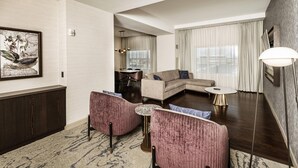 A modern hotel room with a sofa, two armchairs, a small round table, and a large window with curtains.