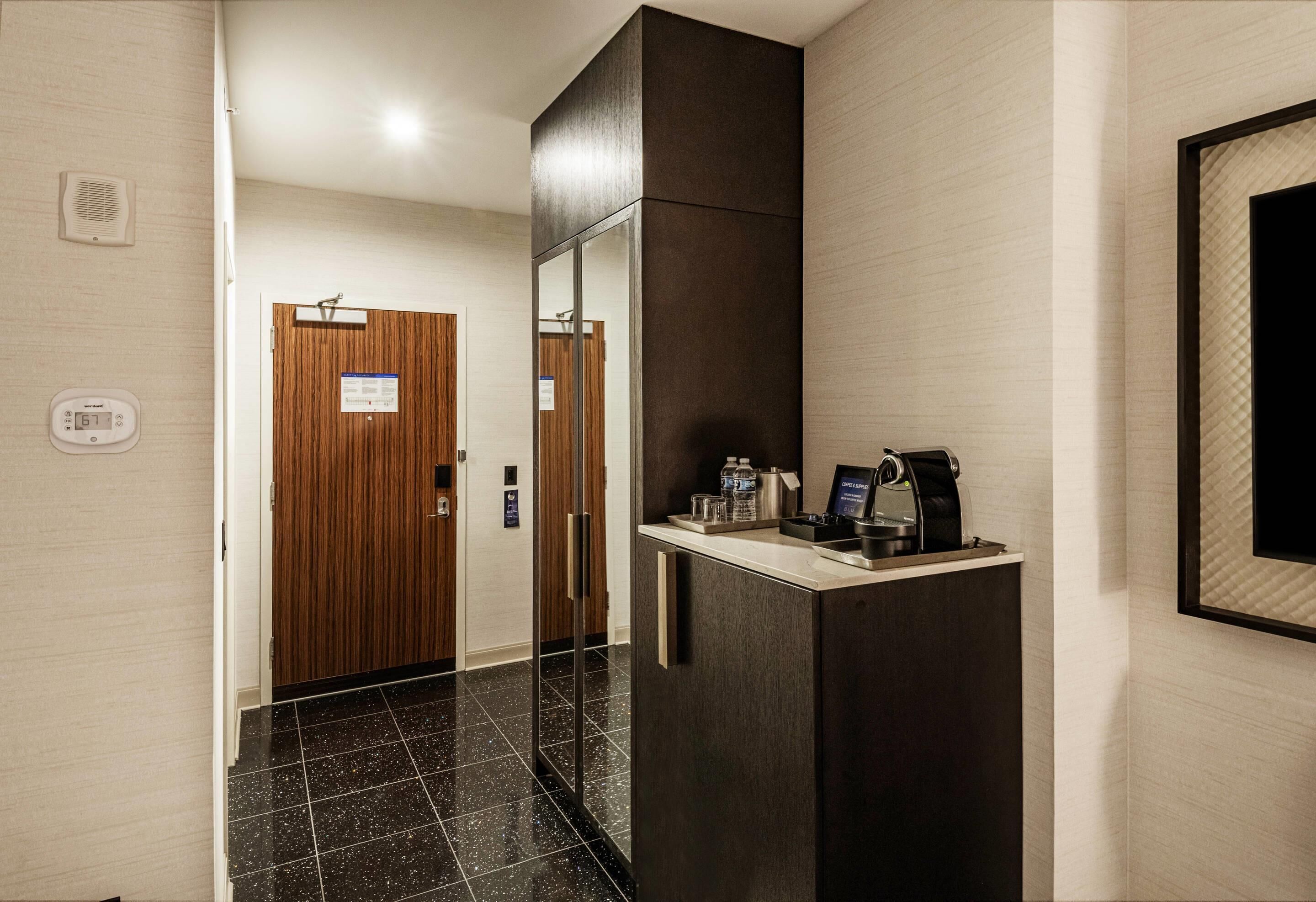 Un pasillo de hotel con una puerta de madera, un termostato digital y una estación de café con cafetera y botellas de agua.