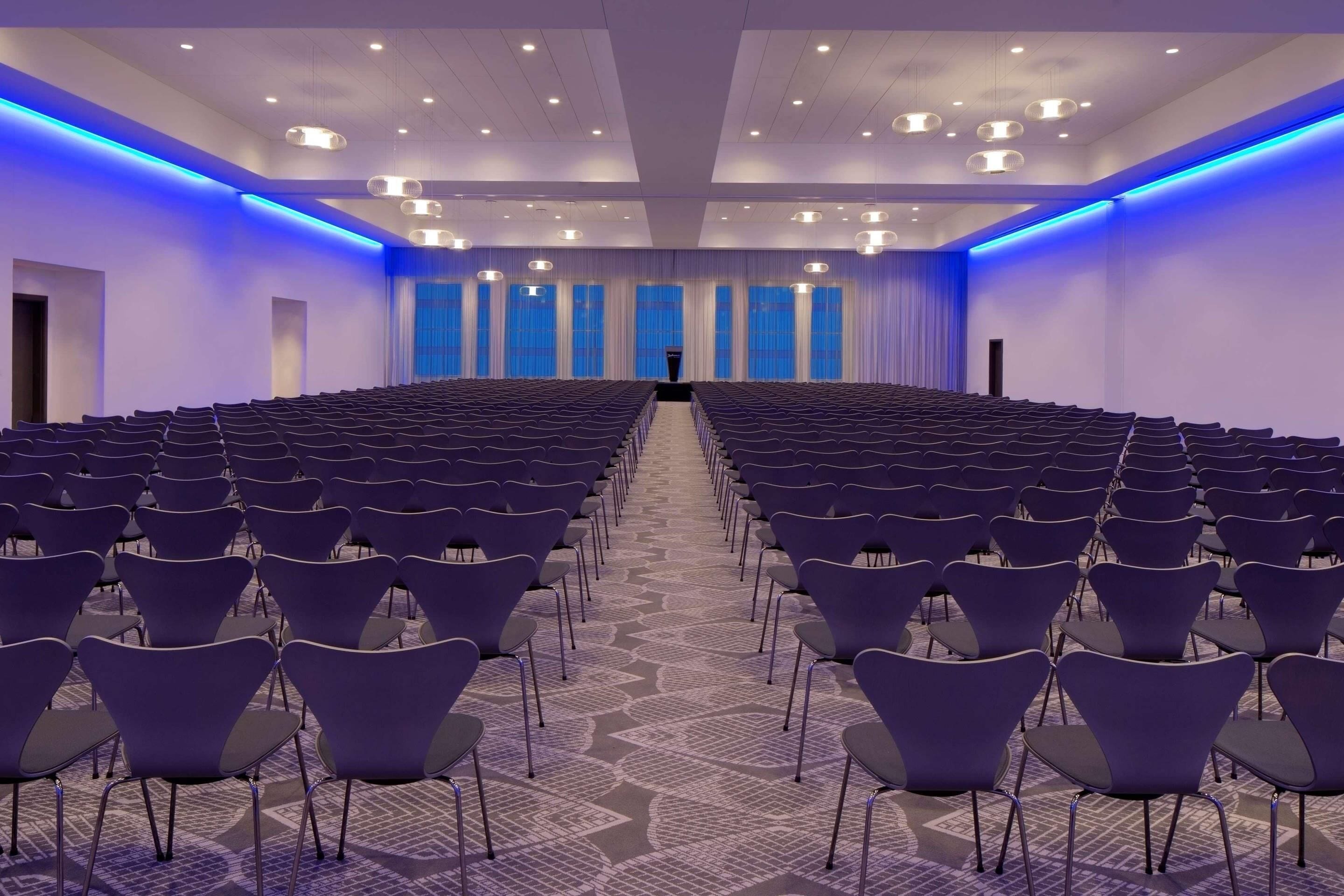 Un salón de conferencias con filas de sillas, un atril y grandes ventanales con cortinas azules.
