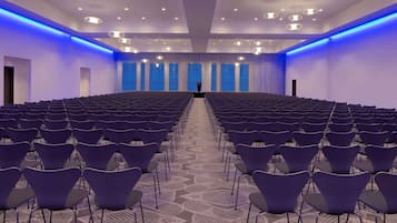 Un salón de conferencias con filas de sillas, un atril y grandes ventanales con cortinas azules.