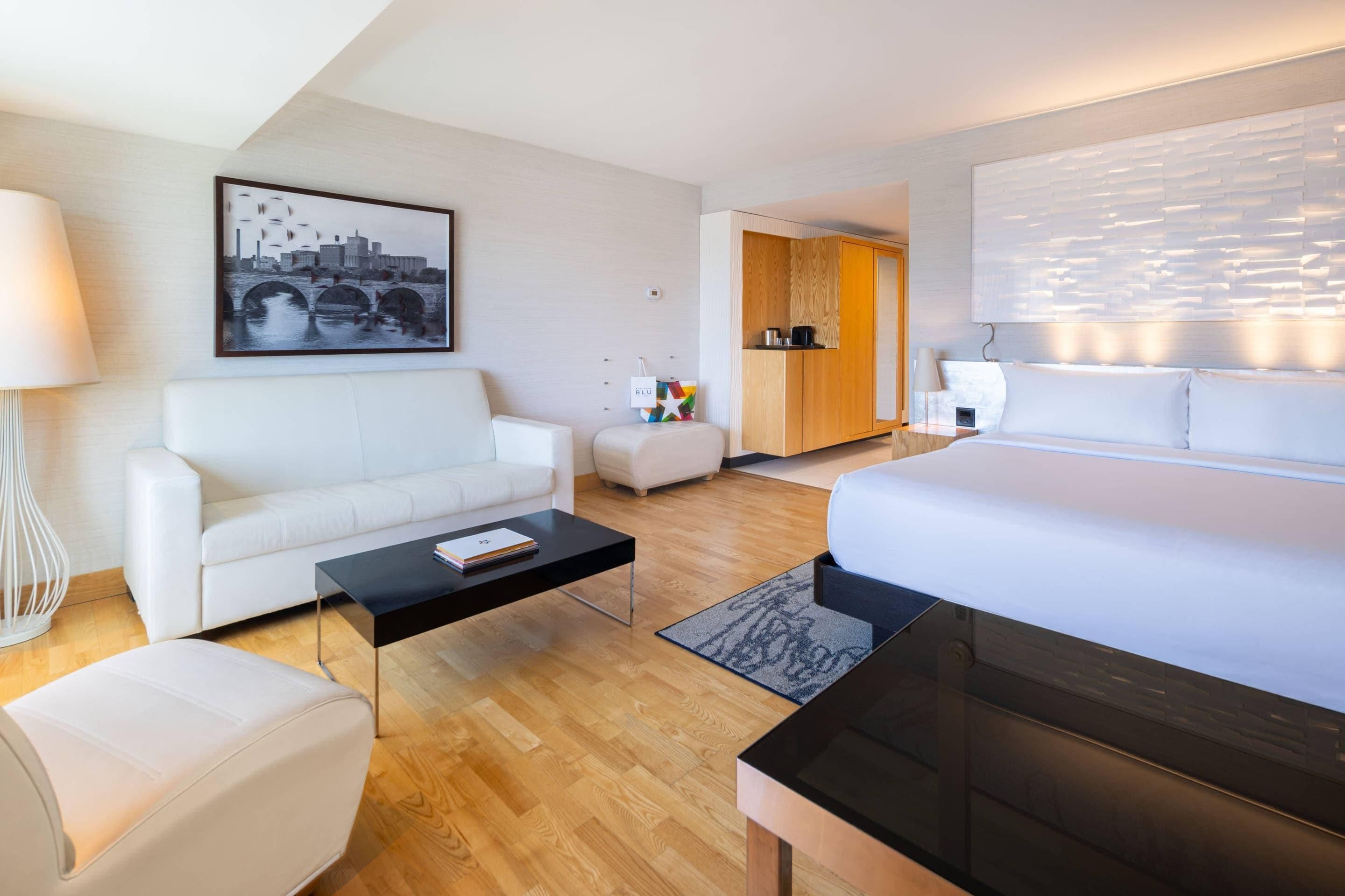 Ett modernt hotellrum med en säng, en soffa, ett soffbord och ett skrivbord. Det finns en tavla på väggen och utsikt över staden.
