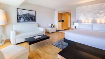 Una habitación de hotel moderna con una cama, un sofá, una mesa de centro y un escritorio. Hay un cuadro en la pared y vistas a la ciudad.