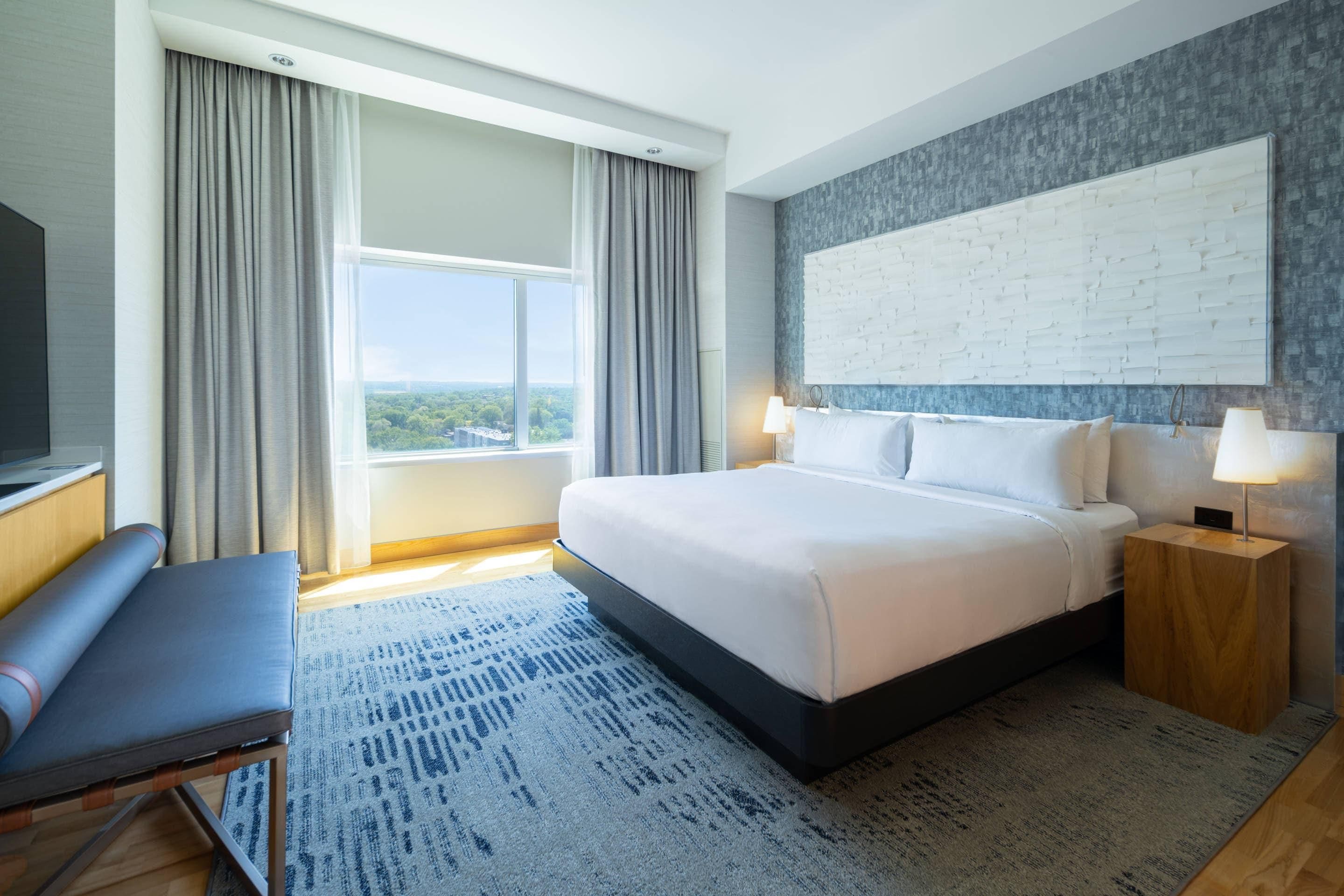A hotel room with a large bed, a desk, a chair, a TV, and a window with curtains.