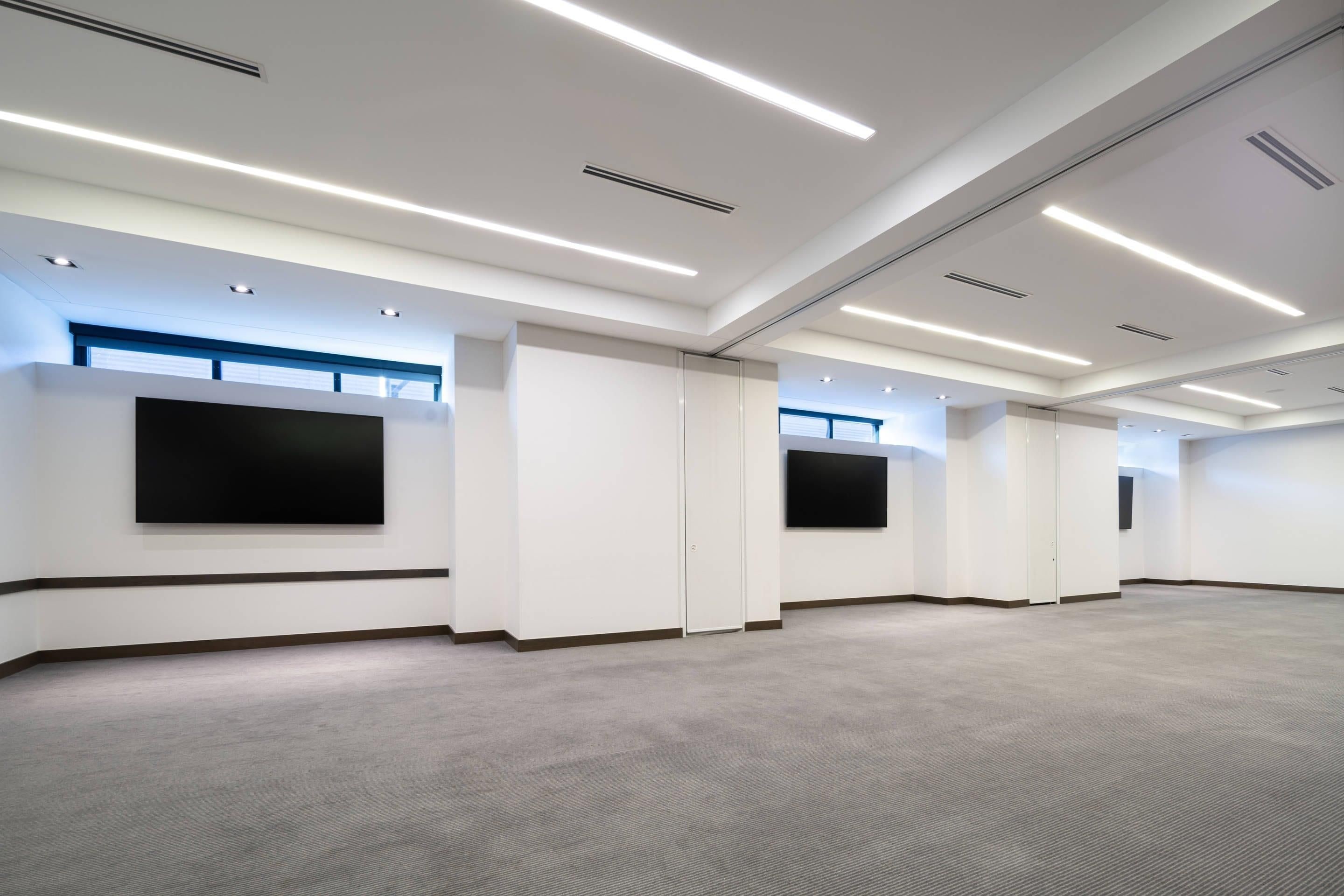 A large, empty conference room with a flat-screen TV mounted on the wall, recessed lighting, and a carpeted floor.