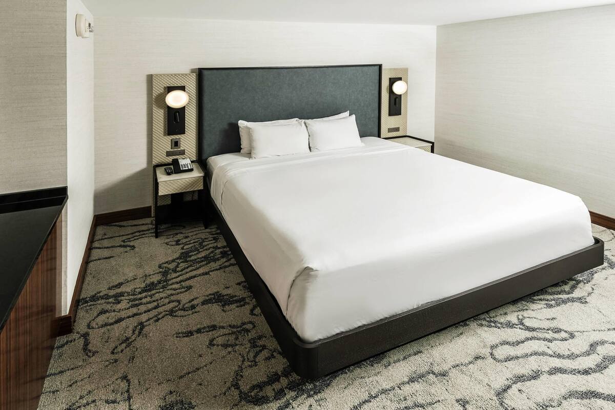 Hotellihuone, jossa on suuri sänky, kaksi yöpöytää valaisimineen ja kuviollinen matto.