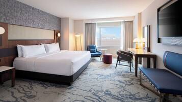 A hotel room with a large bed, a desk, a chair, a TV, and a view of the city.