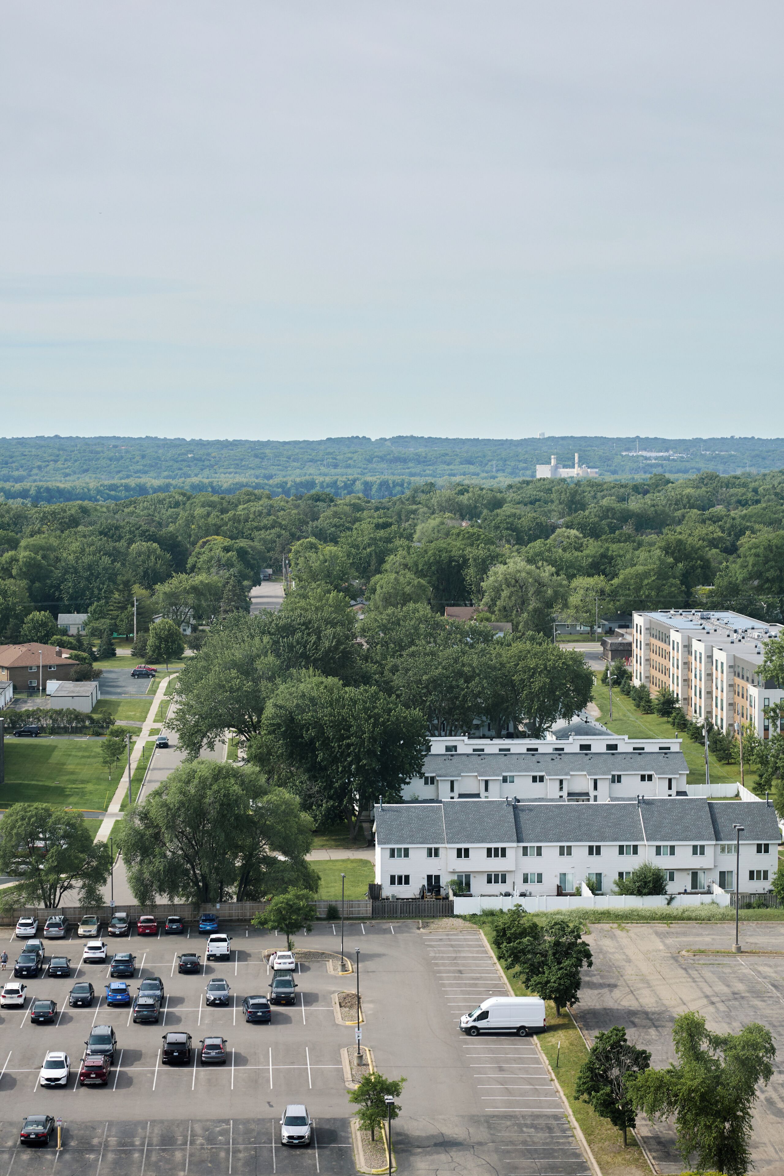 A parking lot with multiple cars and a white van, surrounded by greenery and buildings.