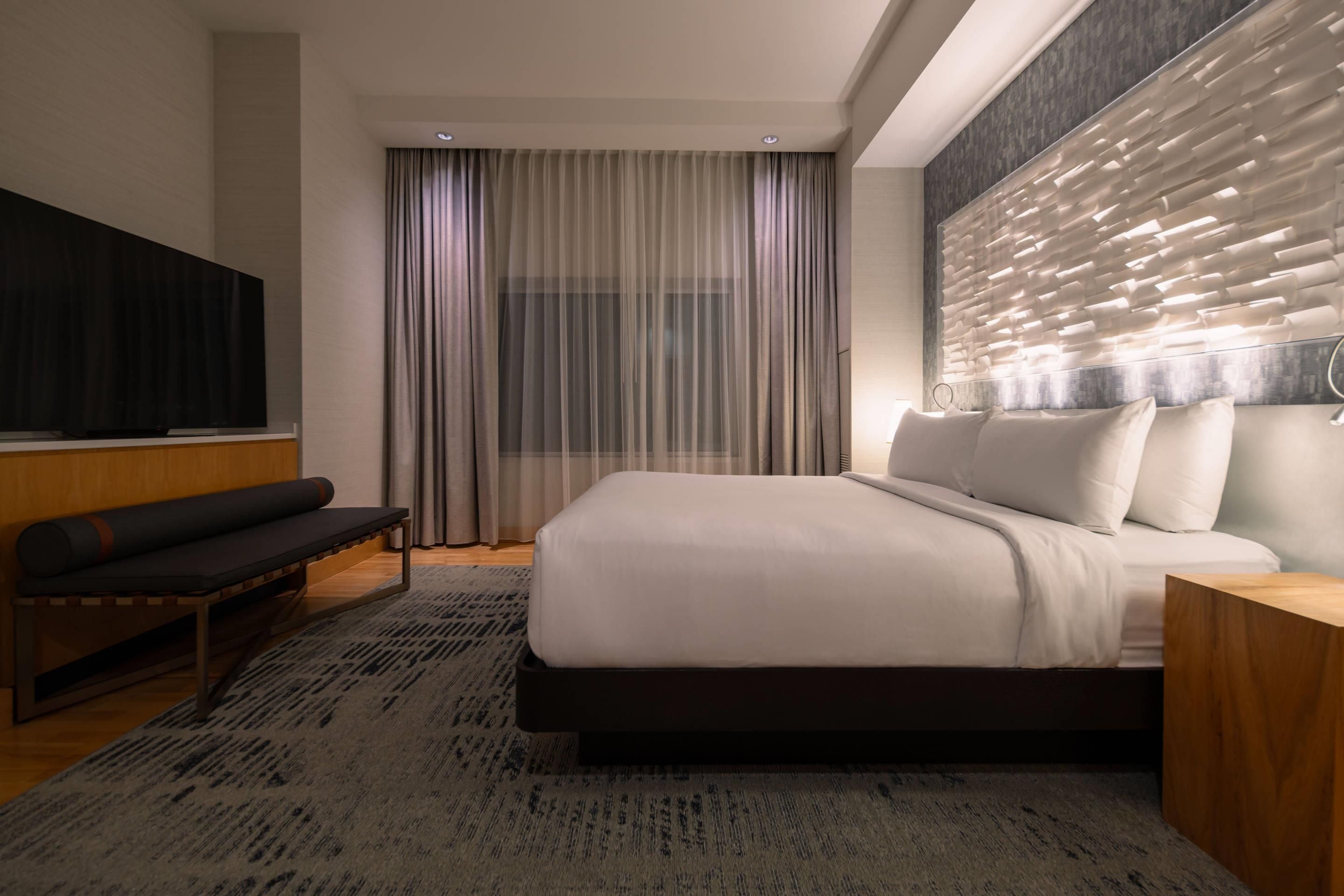 A modern hotel room with a large bed, a bench, a TV, and a decorative wall panel.