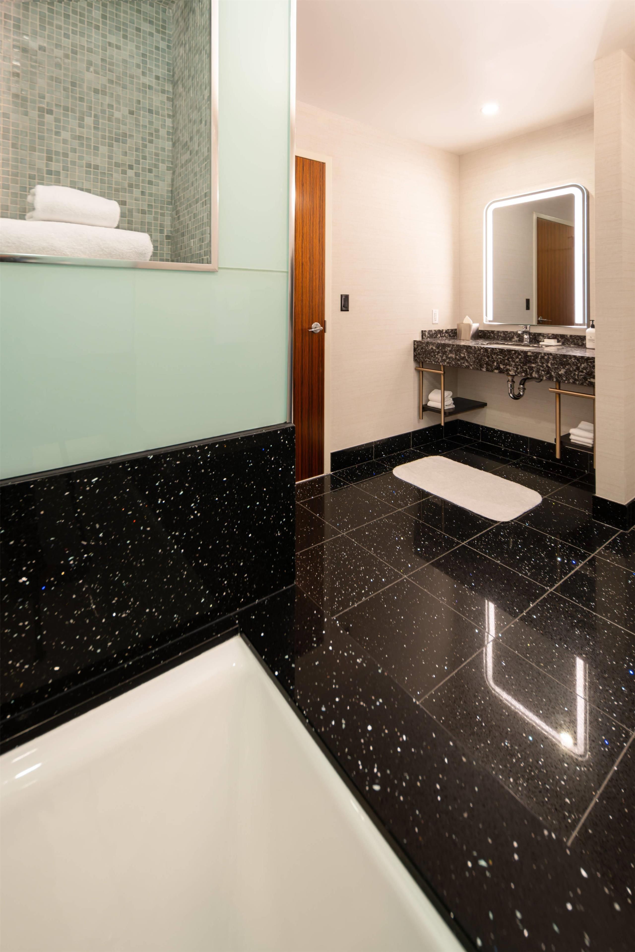 A modern bathroom with a black speckled floor, a white bathtub, and a vanity with a mirror.