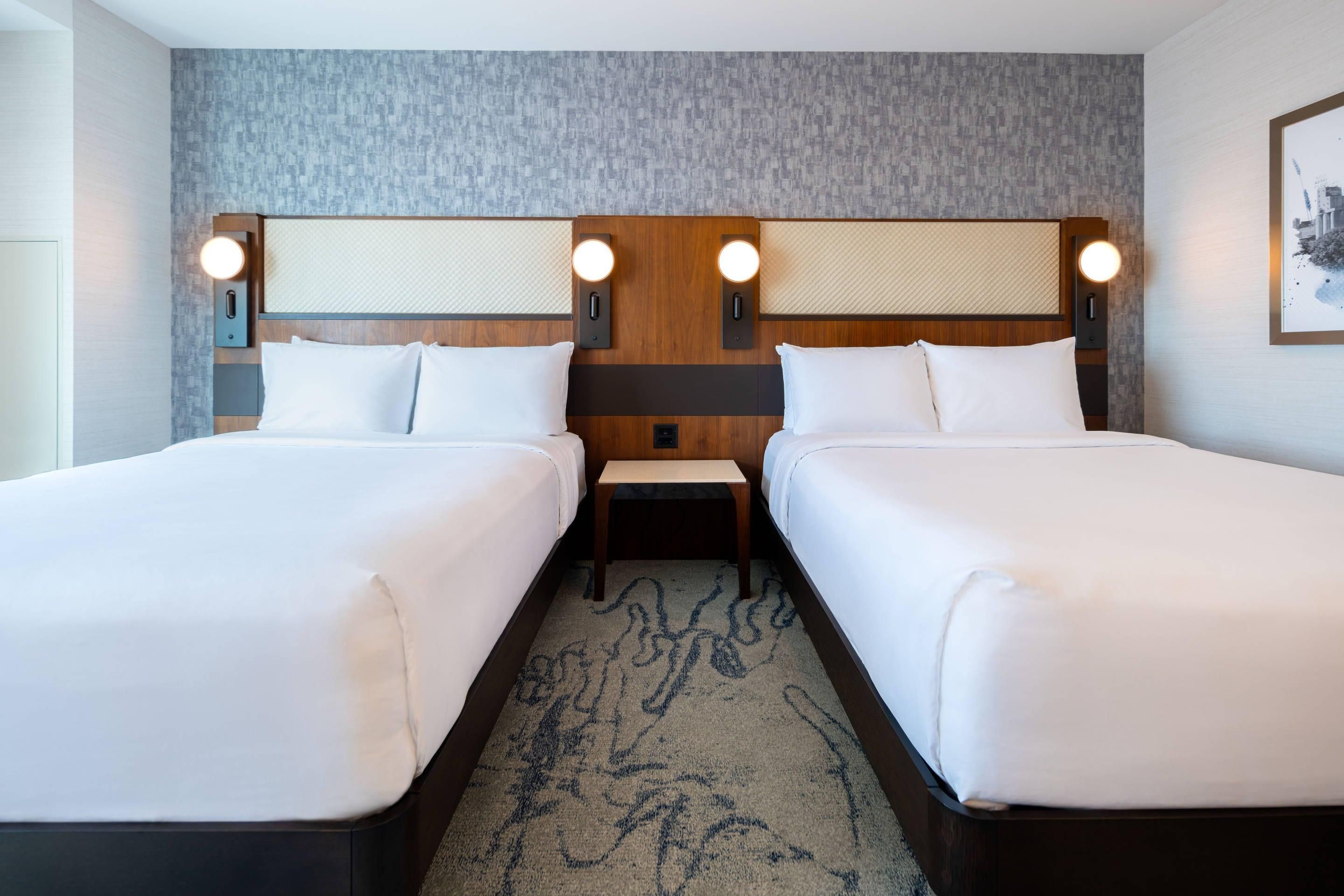 A hotel room with two beds, a small bedside table, a wall-mounted light fixture, and a patterned carpet.