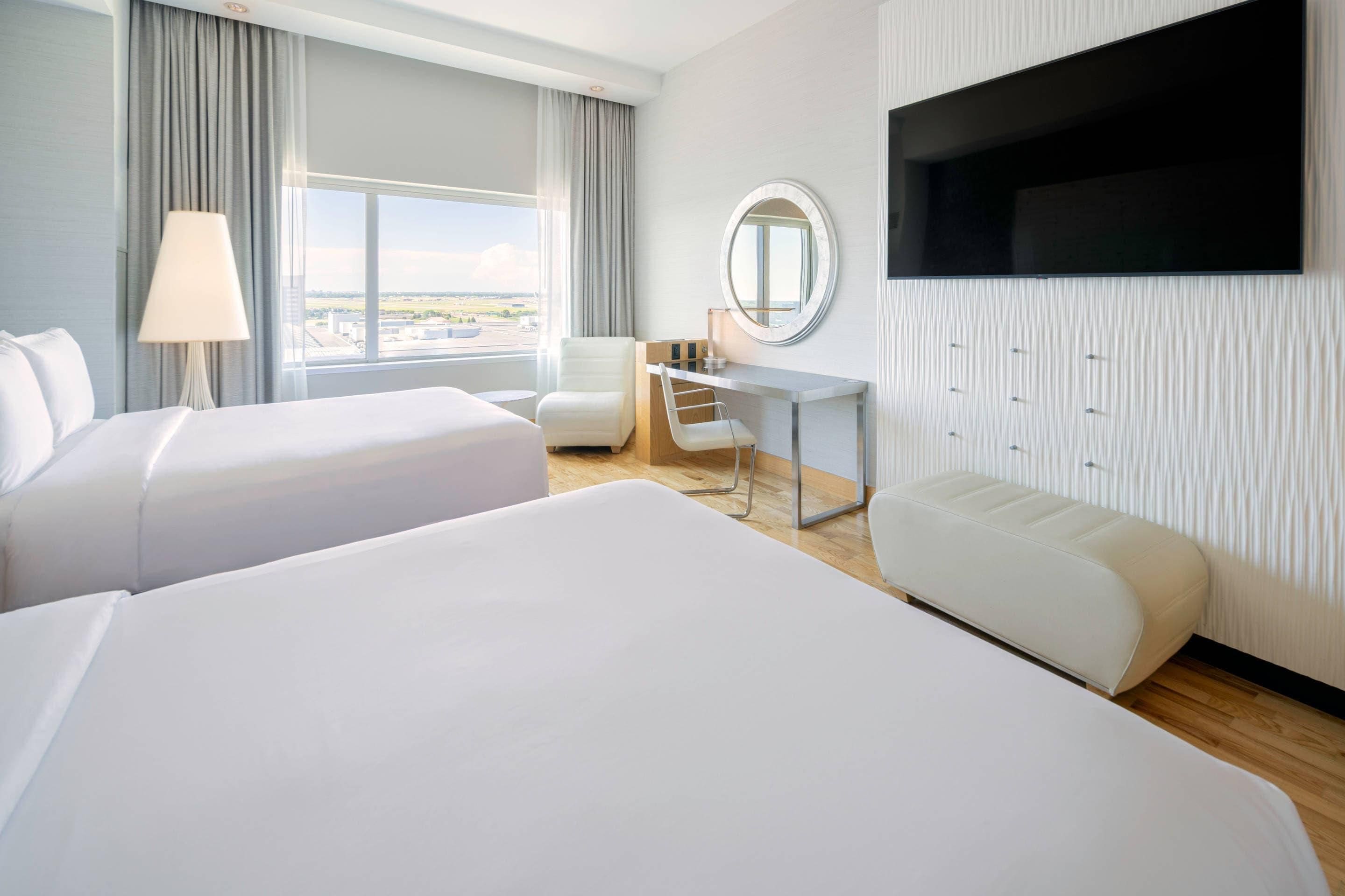 A hotel room with two beds, a desk, a chair, a TV, and a view of the city.