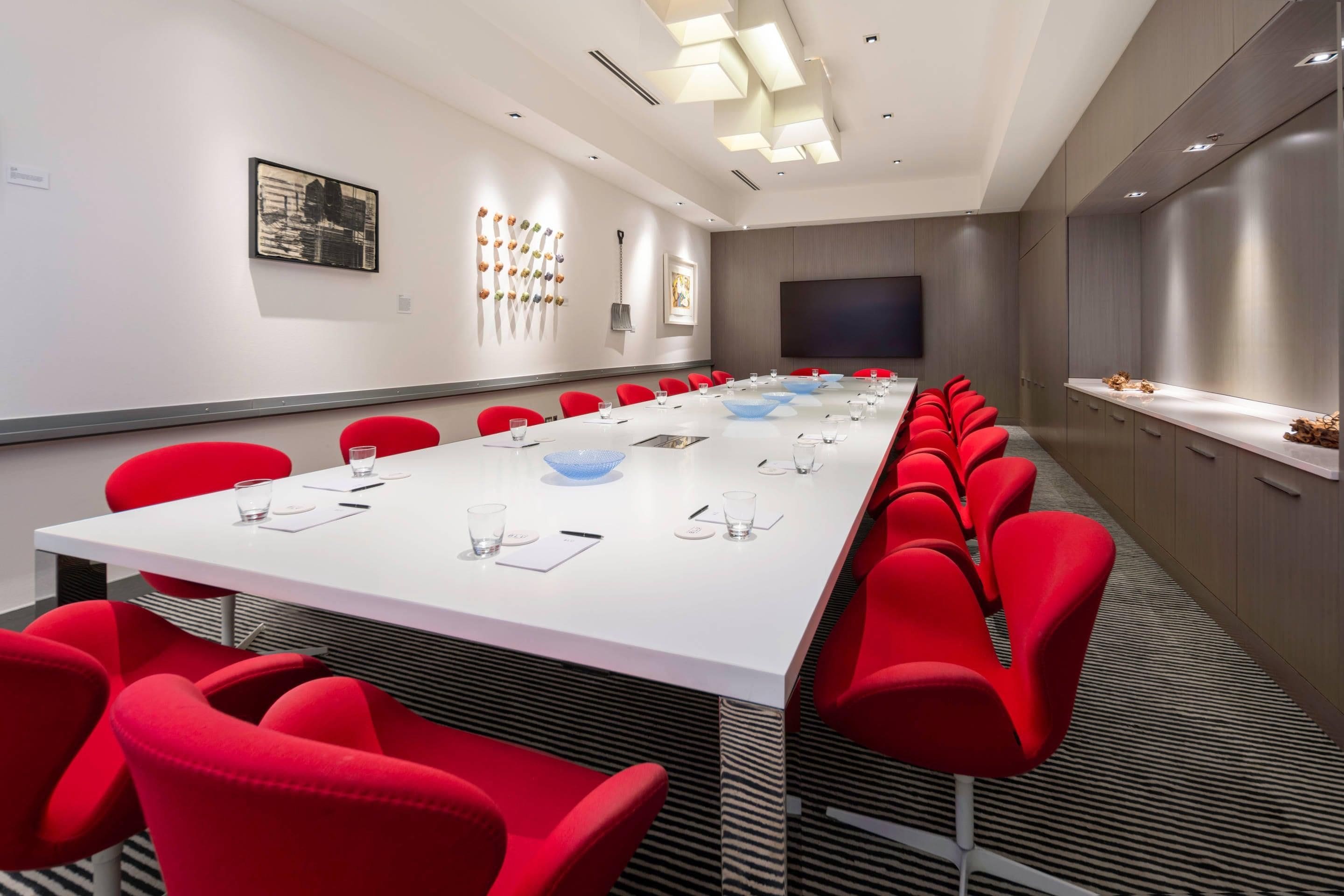 A conference room with a long white table, red chairs, and a large screen.