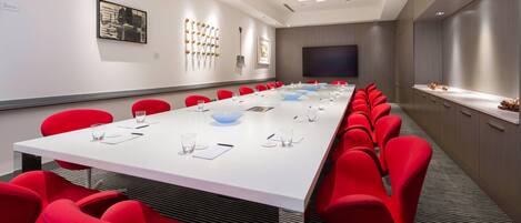 A conference room with a long white table, red chairs, and a large screen.