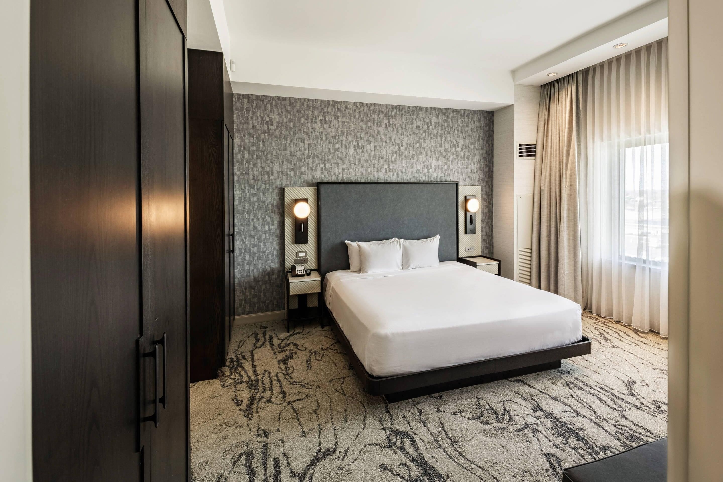 A hotel room with a large bed, dark wood wardrobe, textured wall, and a view of the outdoors through a window.