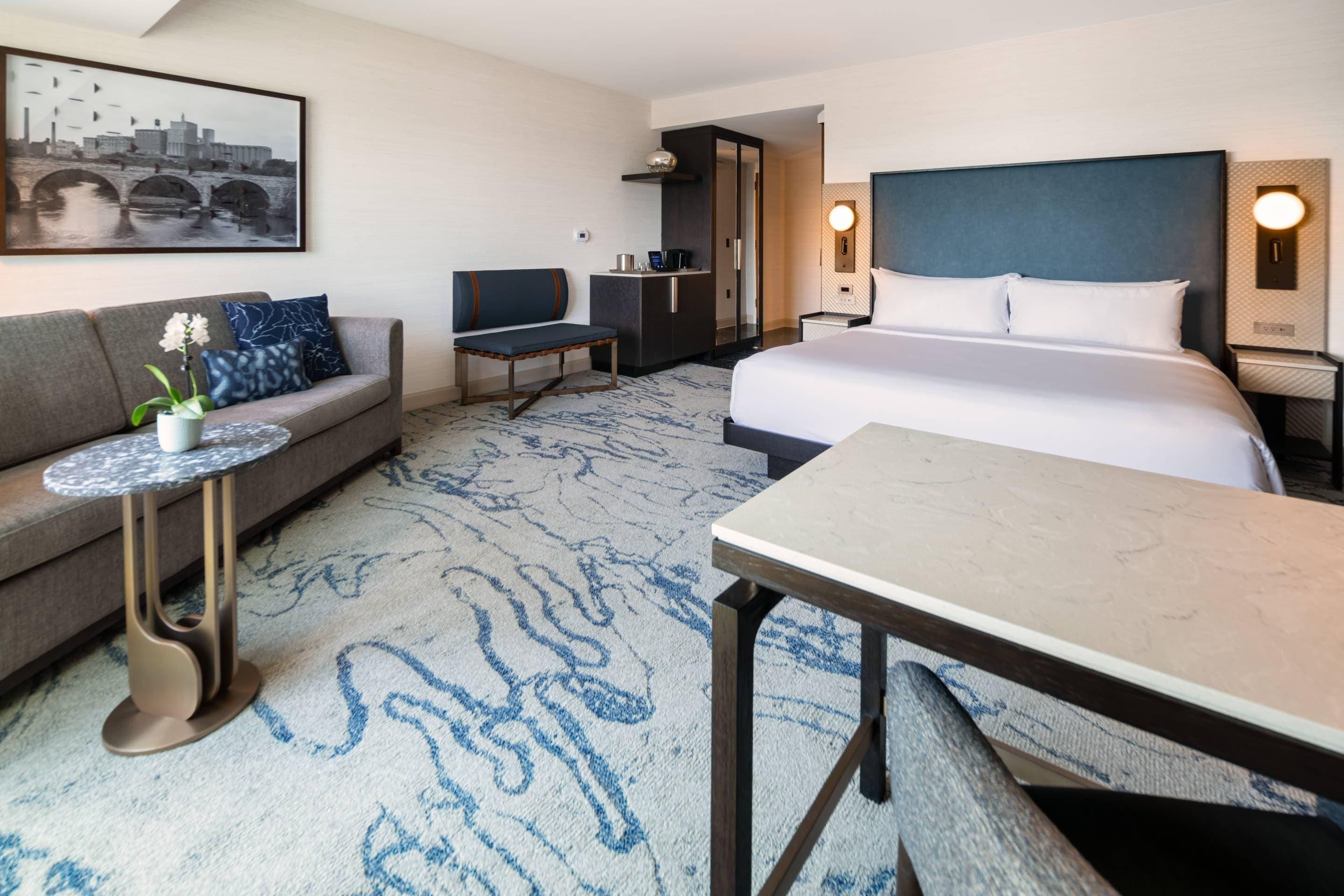 A hotel room with a bed, a sofa, a small table, a desk, and a chair. There is a painting on the wall and a window with a view of a bridge.