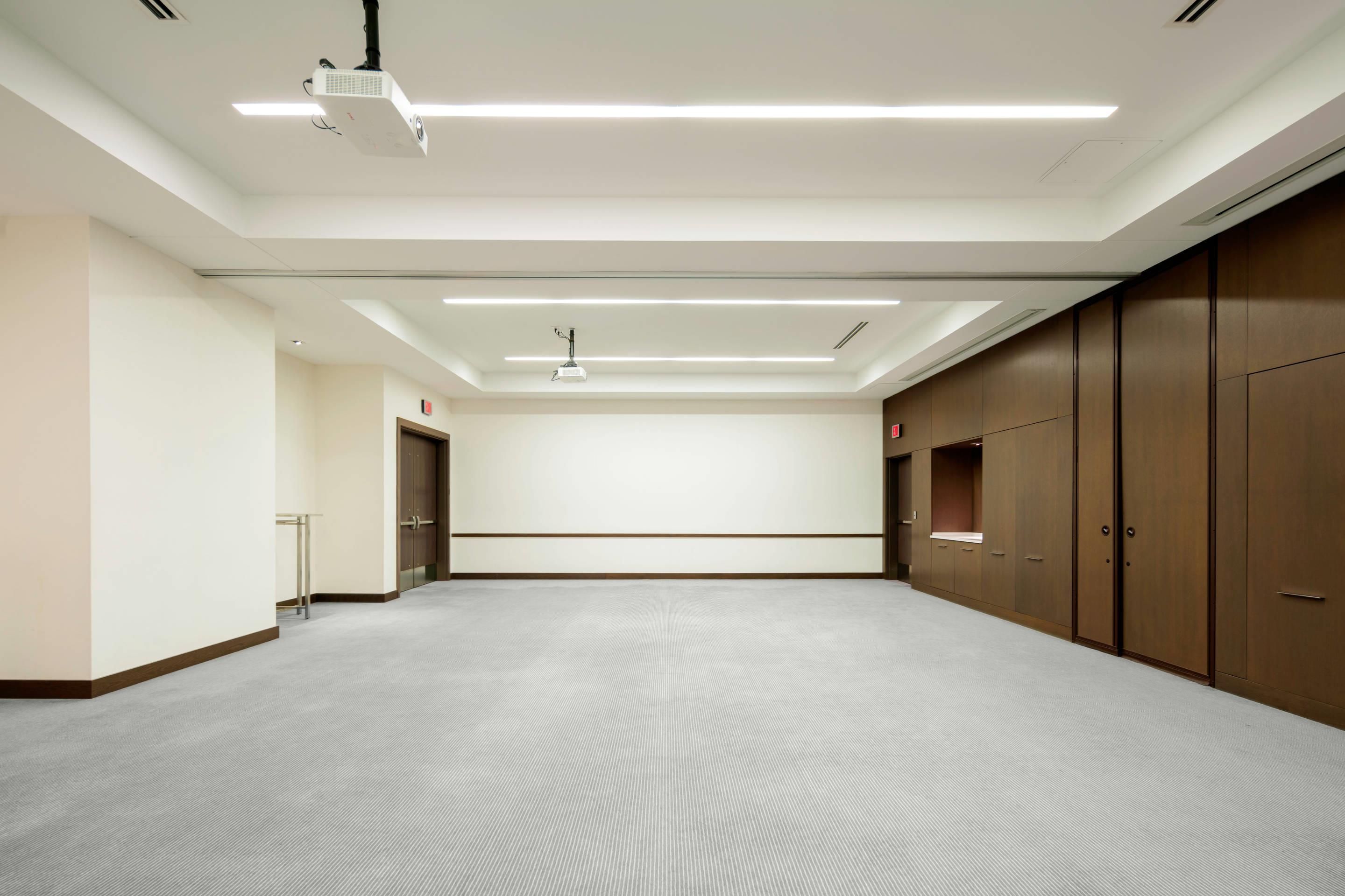 Un salón de conferencias con proyector, luces de techo y piso alfombrado.