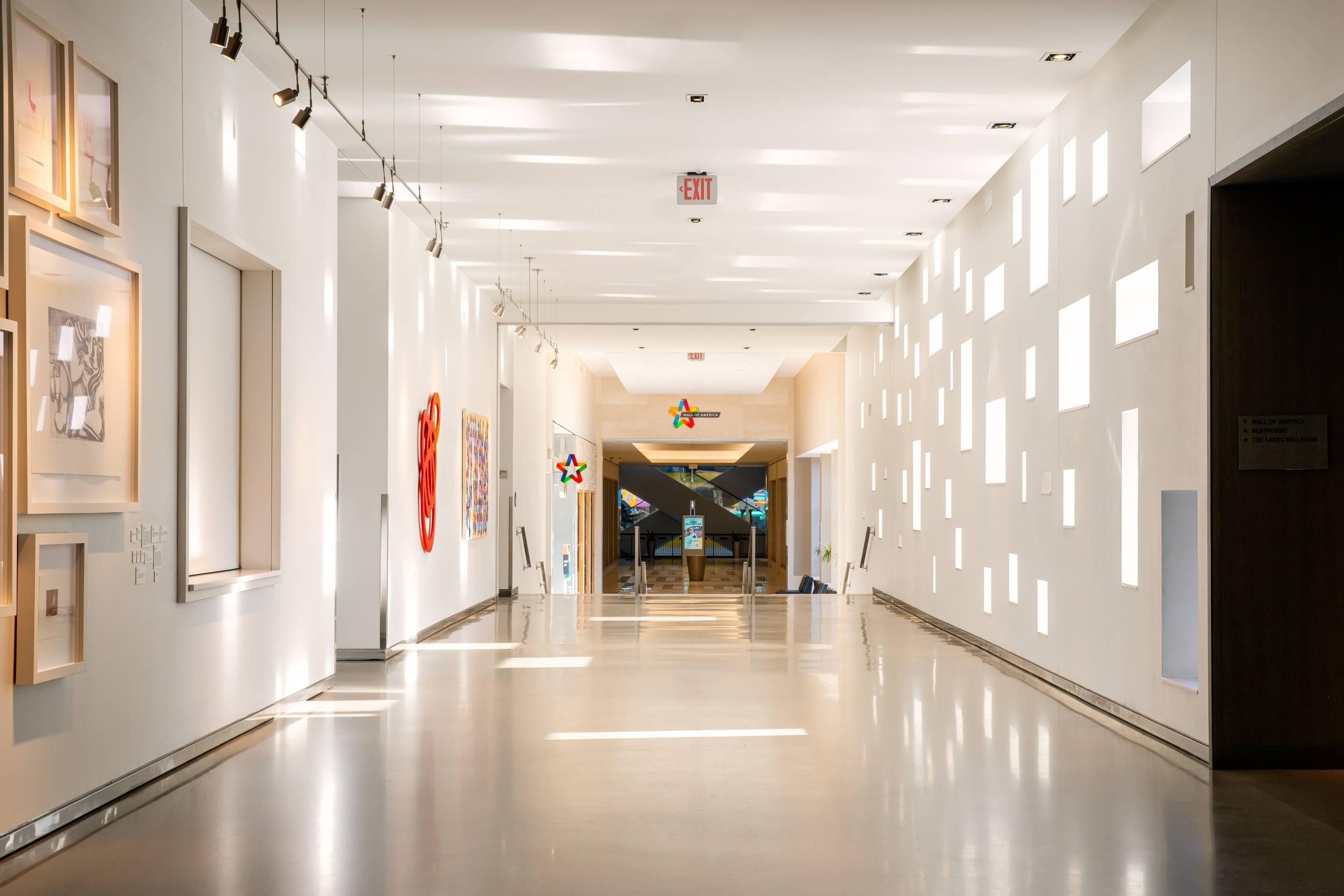 A spacious corridor with a polished floor, directional signage, and framed artwork on the walls.
