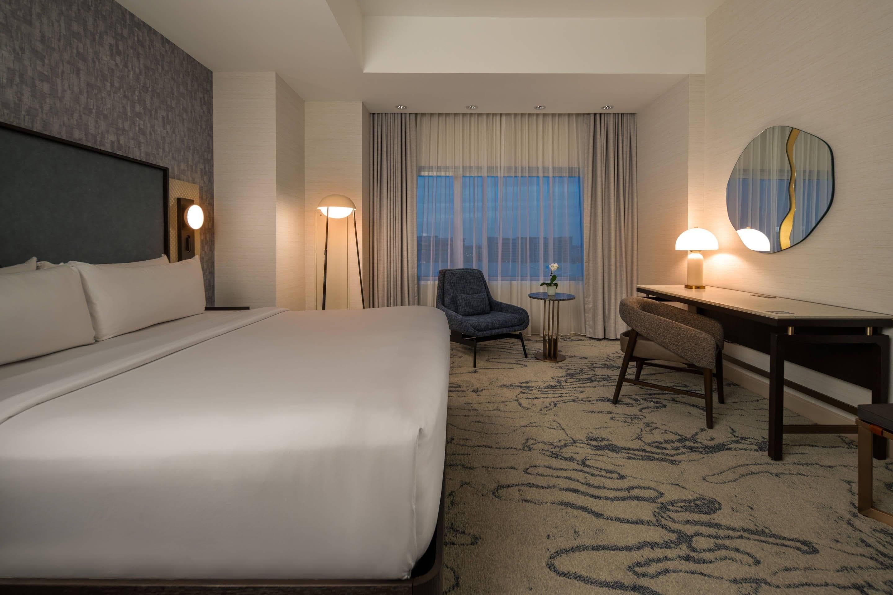 A hotel room with a large bed, a desk, a chair, a small table with a lamp, and a large mirror on the wall.