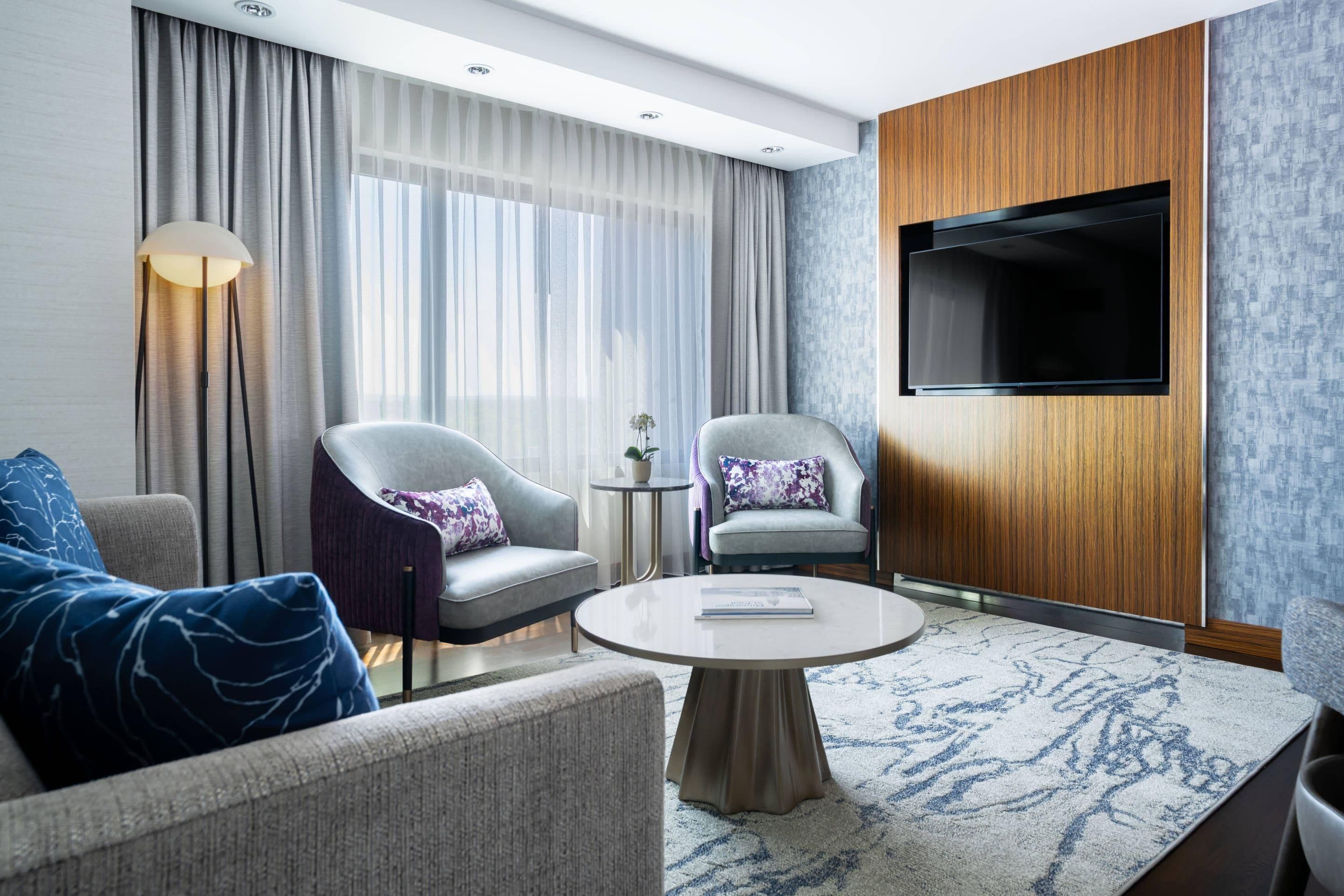 A modern hotel room with two armchairs, a sofa, a round coffee table, a flat-screen TV, and a large window with sheer curtains.