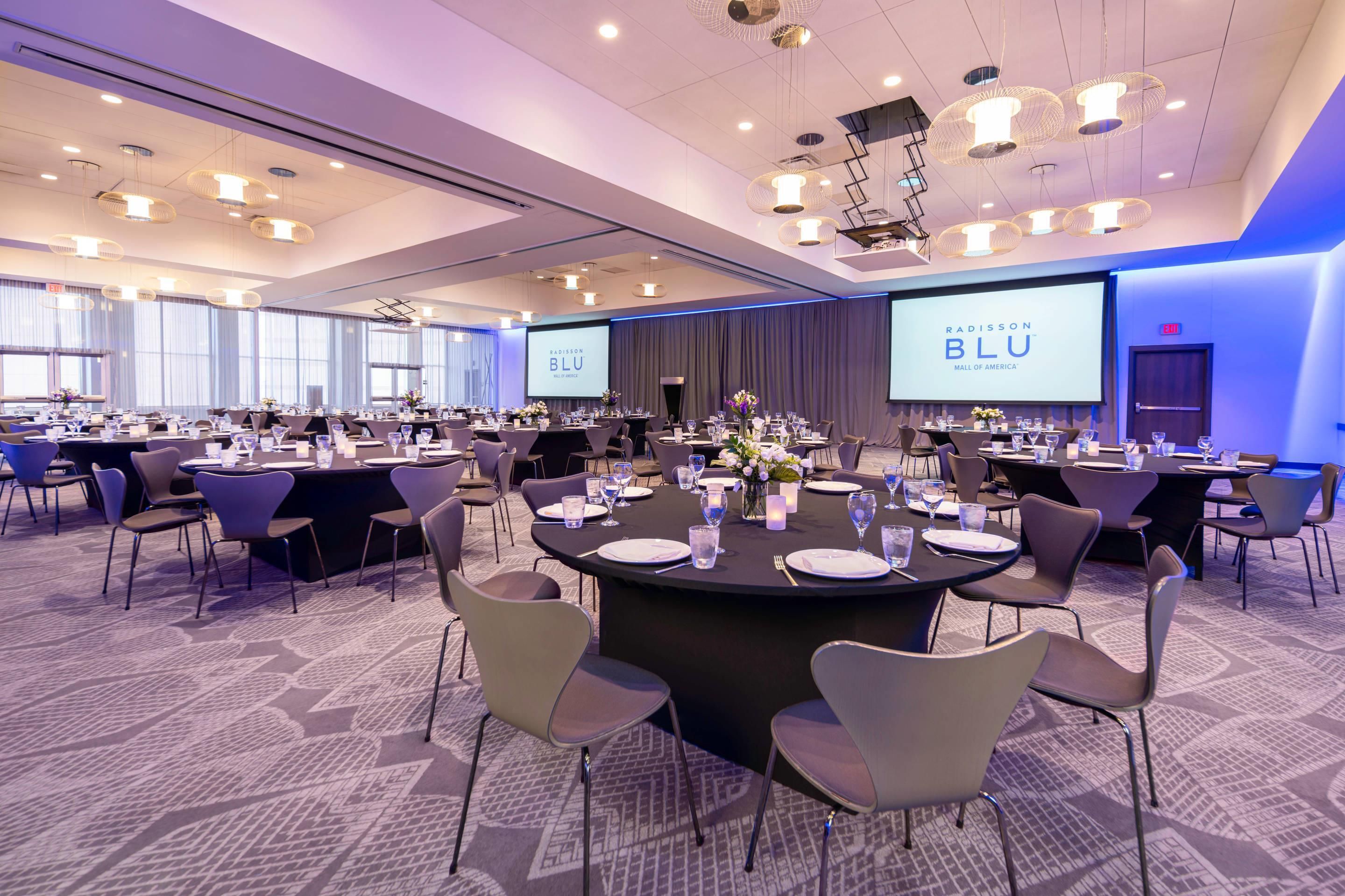 A conference room with round tables, chairs, and a large screen displaying 'RADISSON BLU MALL OF AMERICA'.