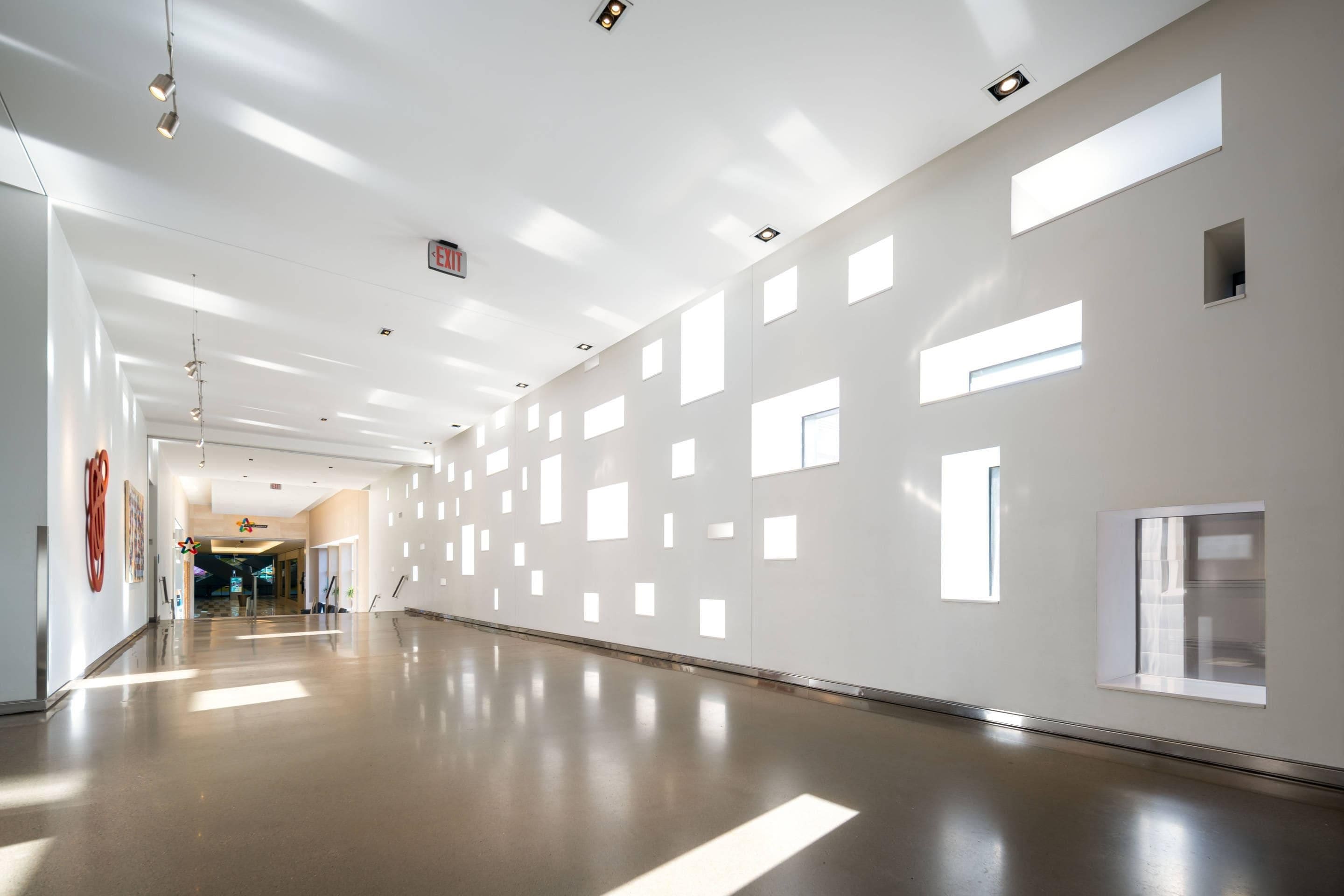 A spacious corridor with a polished floor, recessed lighting, and a series of rectangular windows on the walls.