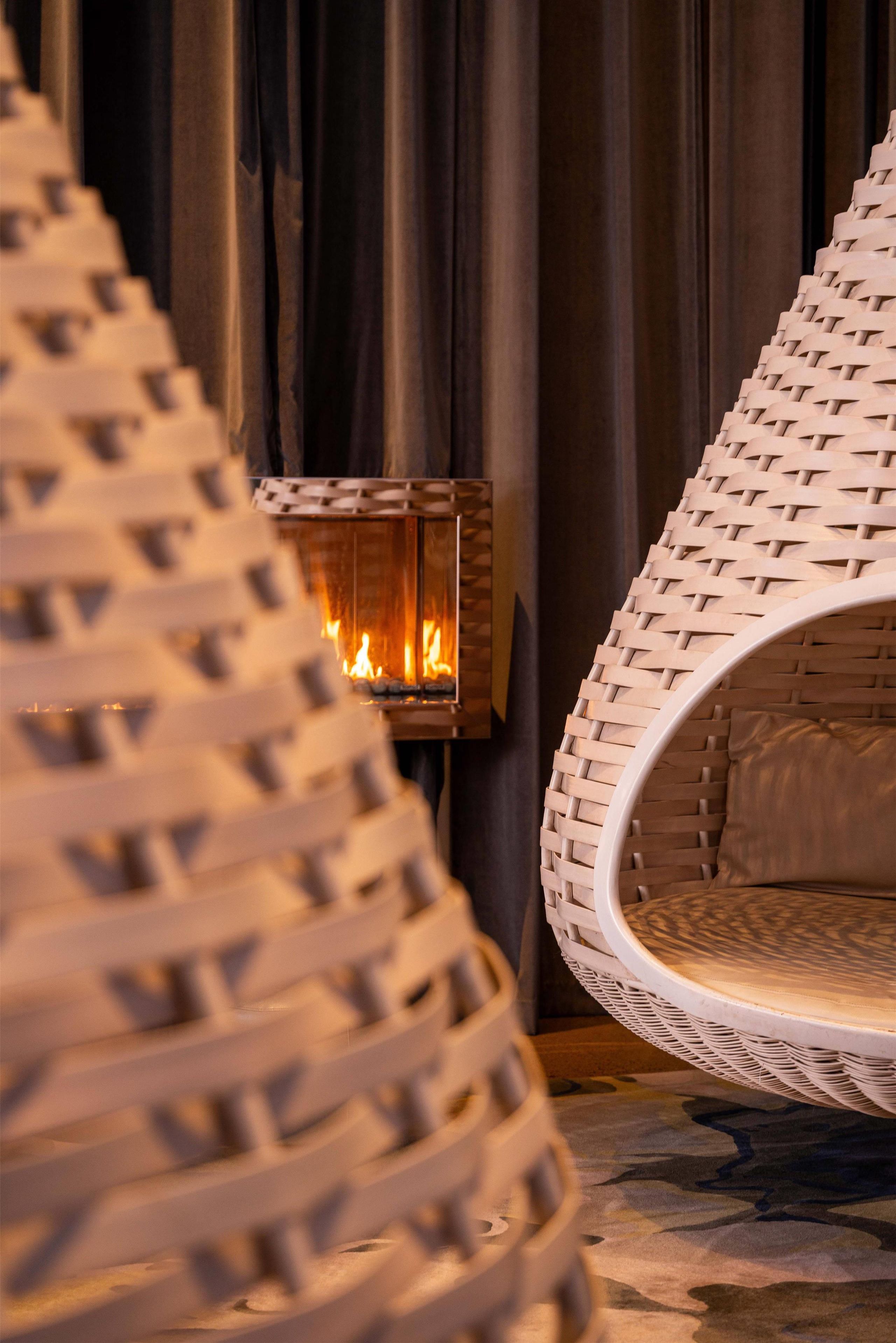Una silla de mimbre con asiento y respaldo circulares, ubicada frente a una chimenea con llamas visibles.