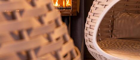 Una silla de mimbre con asiento y respaldo circulares, ubicada frente a una chimenea con llamas visibles.