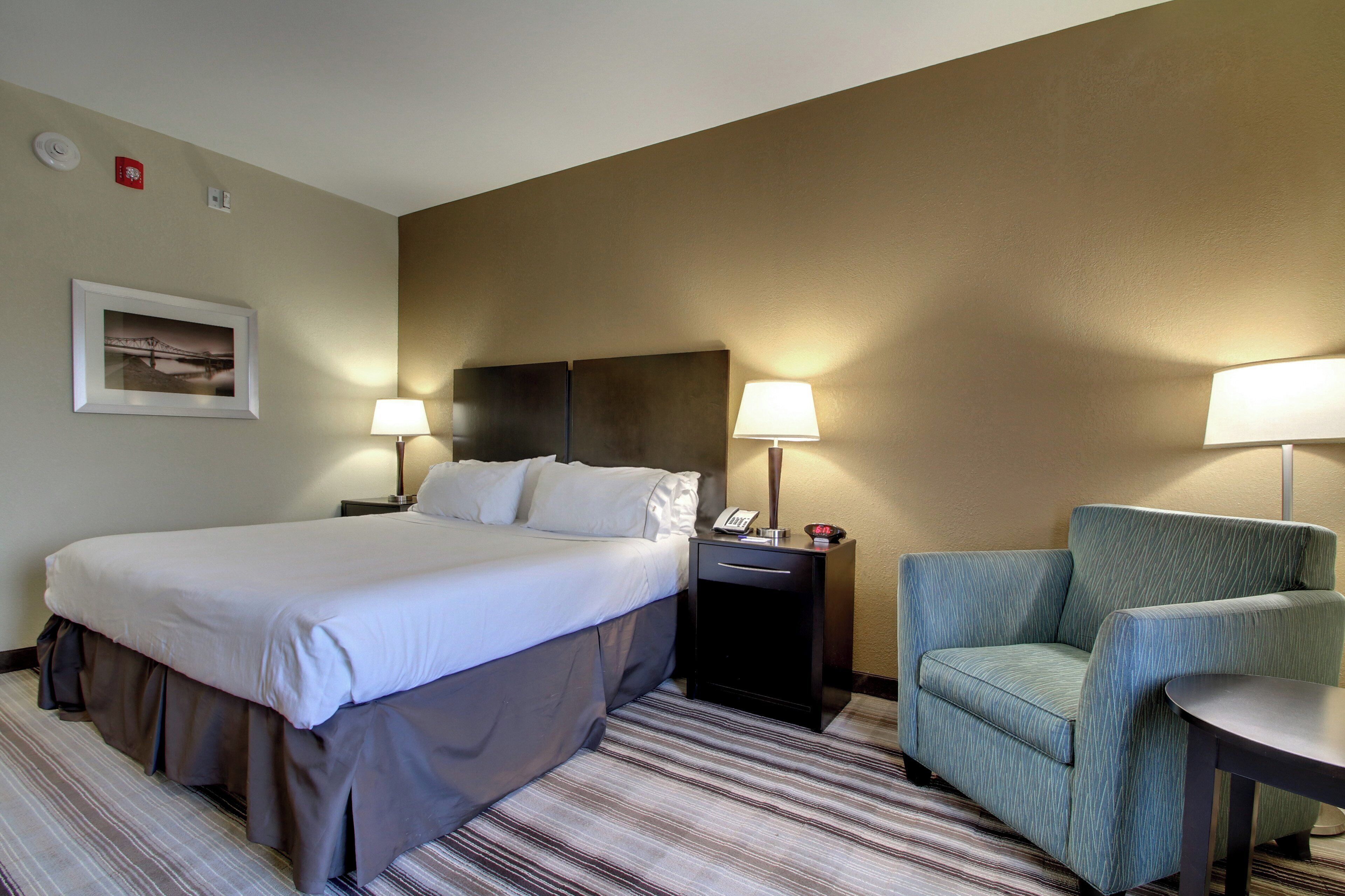 Quarto de hotel com uma cama grande, duas mesas de cabeceira com luminárias, uma cadeira e uma pequena mesa.