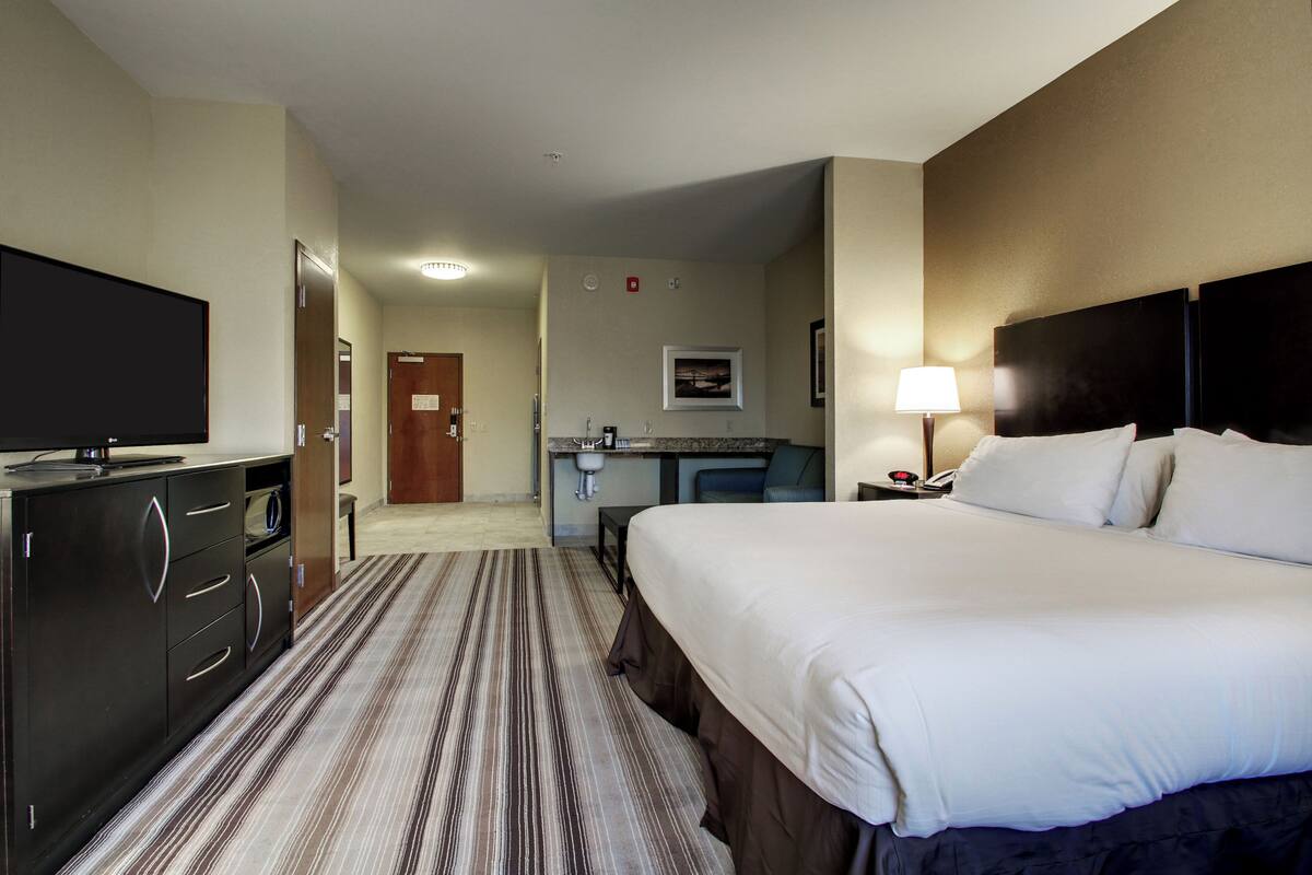 A hotel room with a large bed, a TV on a dresser, a desk with a chair, and a small kitchenette.