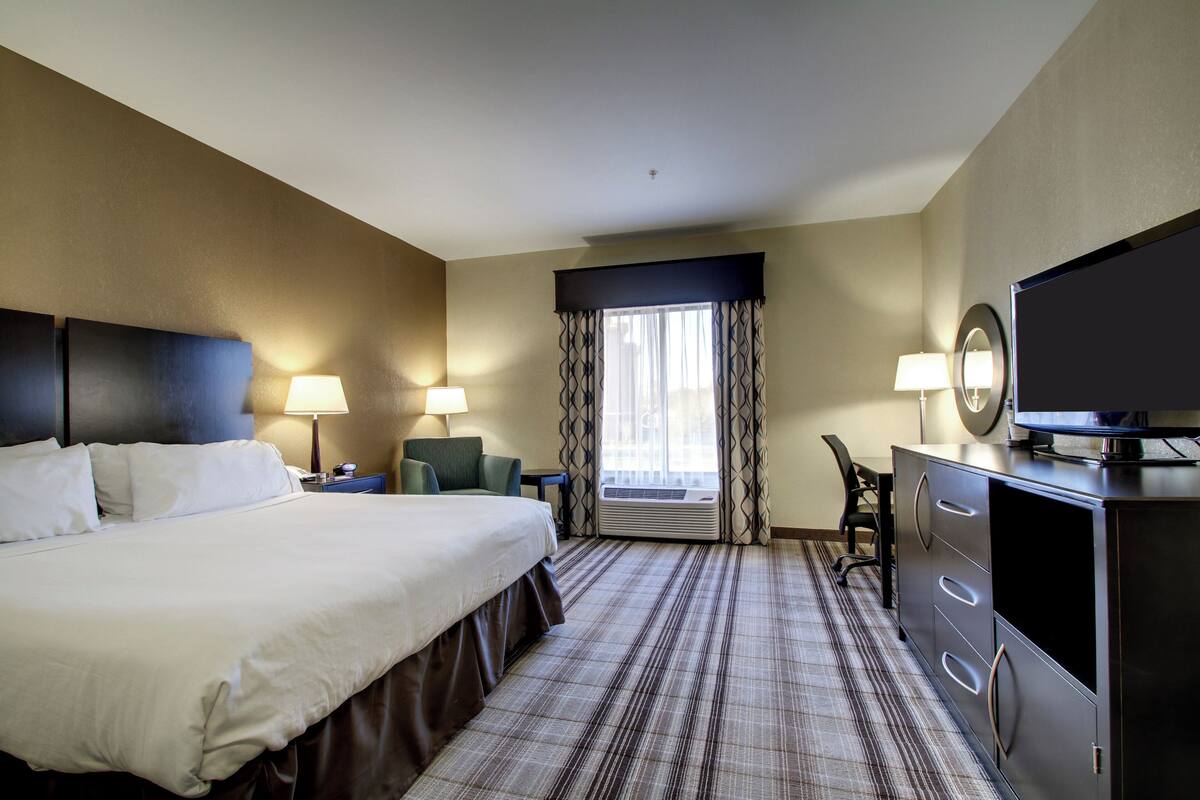 A hotel room with a large bed, two bedside tables with lamps, a desk with a chair, a television, and a window with curtains.