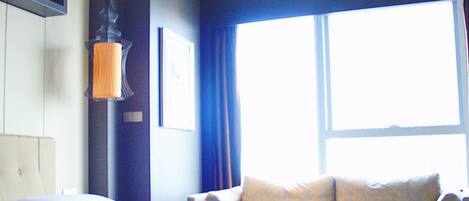 A modern hotel room with a large bed, a seating area with cushions, a wall-mounted shelf, and a large window providing natural light.
