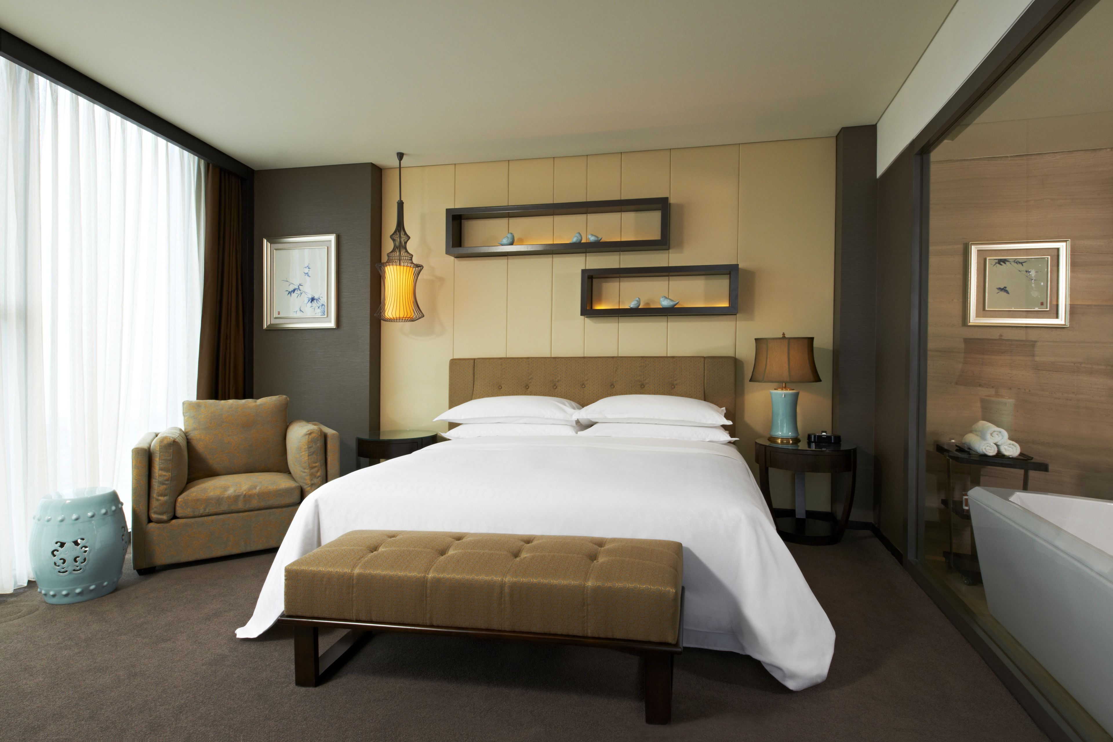 Una habitación de hotel con una cama grande, una silla, un banco, una mesita de noche, una lámpara y una ventana con cortinas.