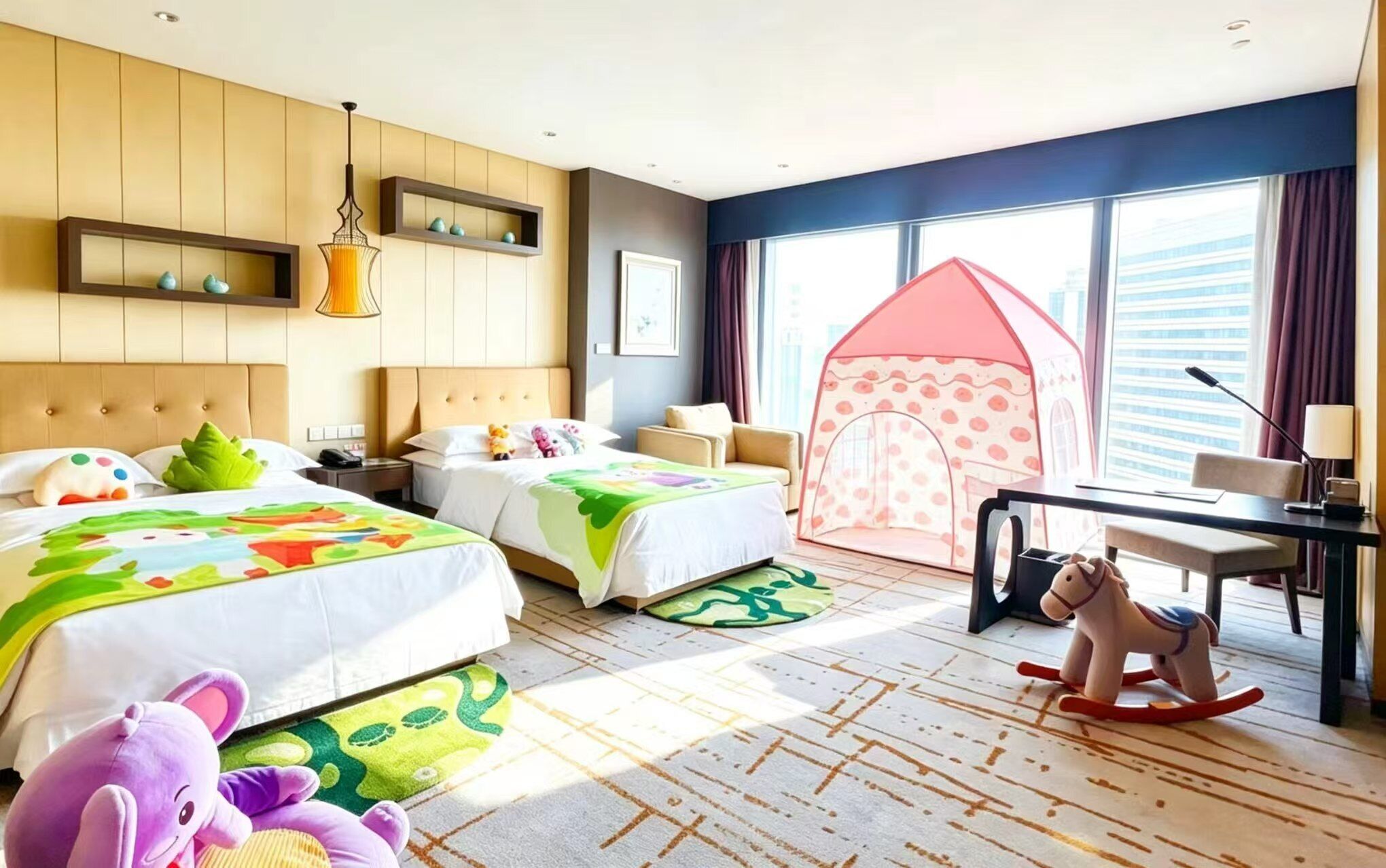 A hotel room with two beds, a desk, a rocking horse, and a play tent.