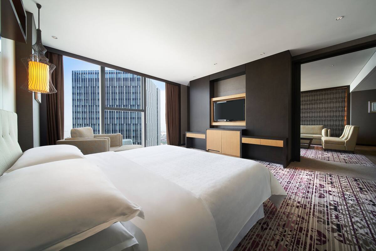 A modern hotel room with a large bed, a TV, a desk, and a view of a cityscape.