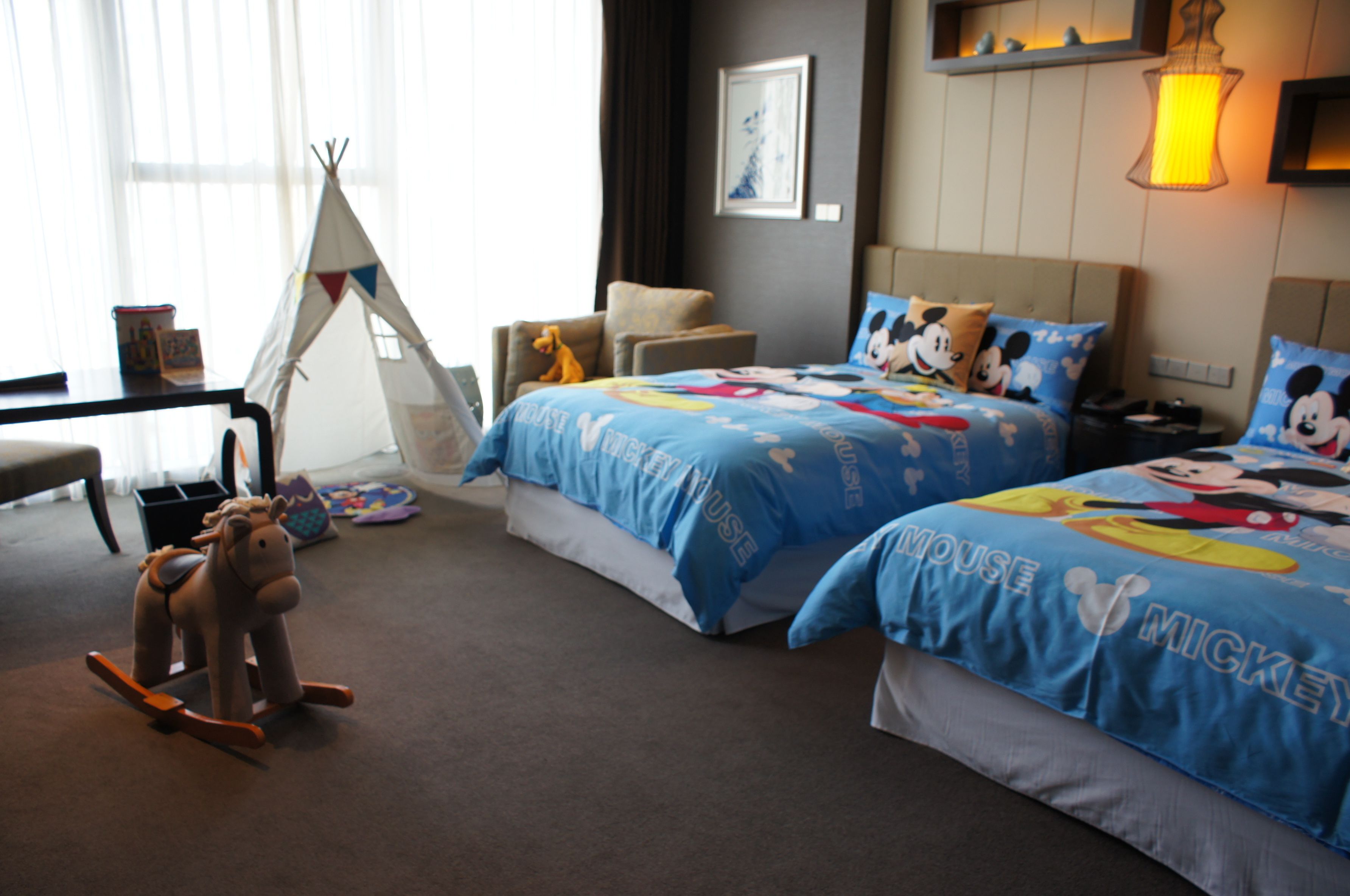 A hotel room with two beds, each with Mickey Mouse themed bedding. There is a rocking horse and a teepee in the room.