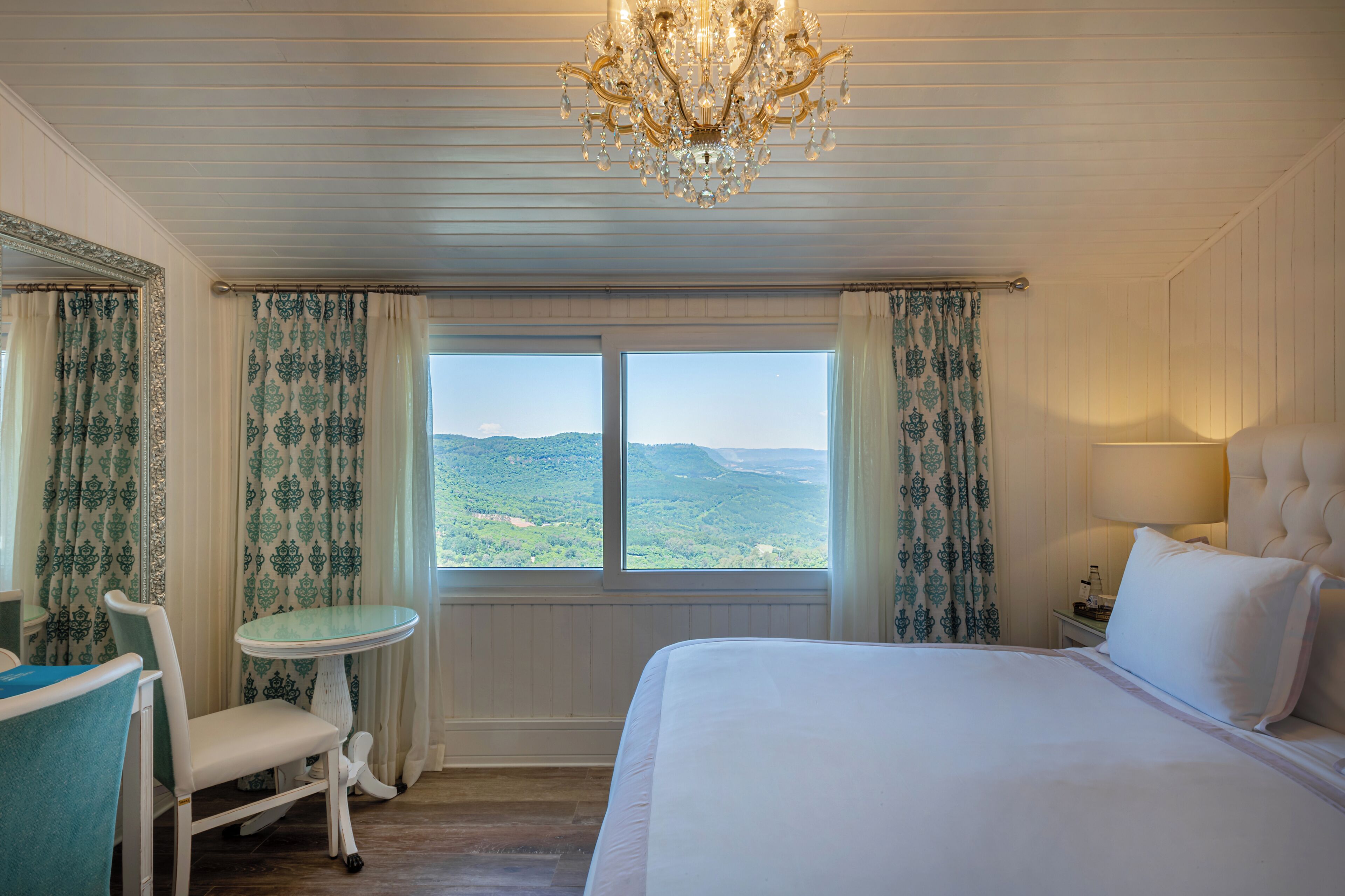 A hotel room with a large bed, a chandelier, a small table, and curtains.