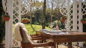 Una mesa de madera dispuesta para comer, con una silla blanca, una estructura de dosel blanca y macetas colgantes.