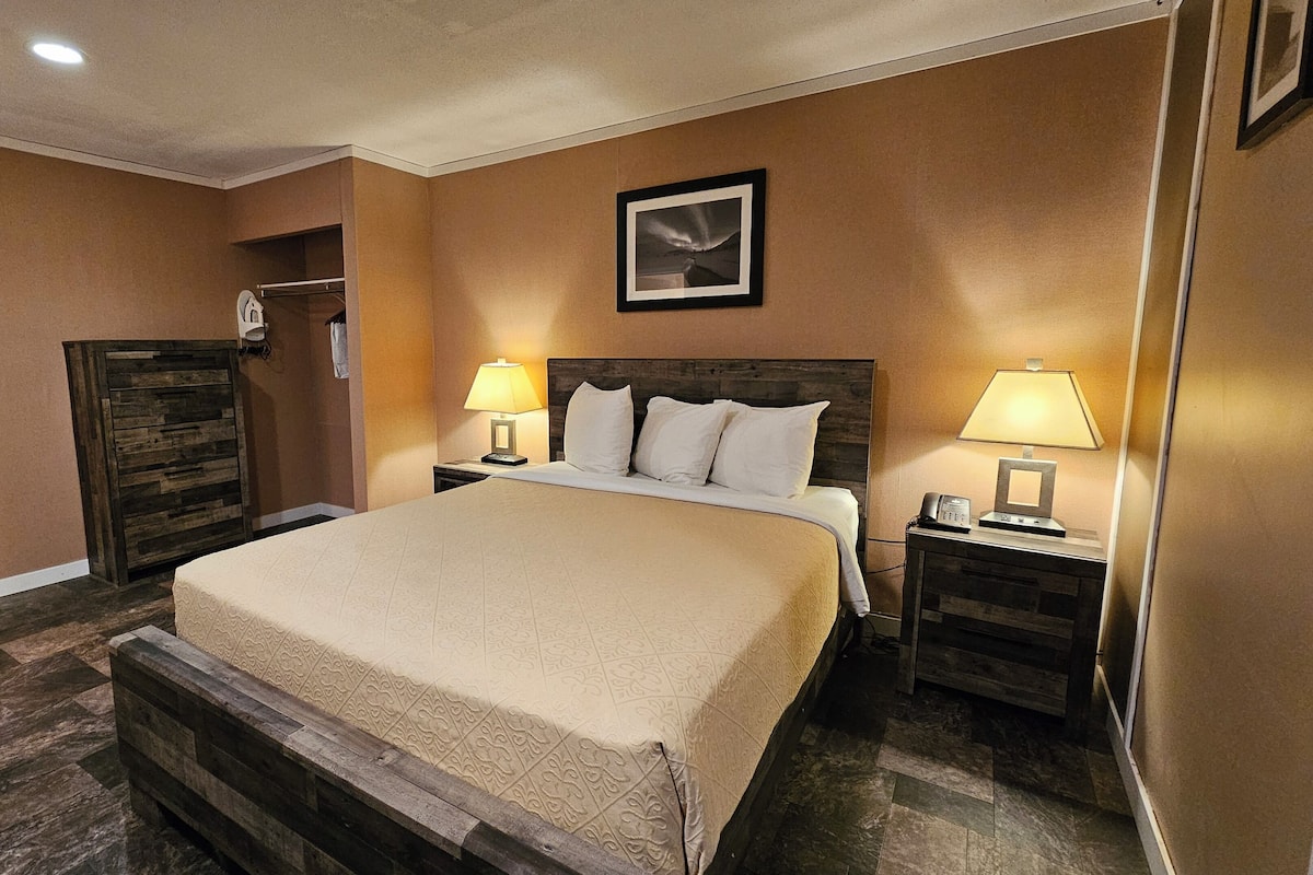 A hotel room with a large bed, two bedside tables with lamps, a wooden bench, a wardrobe, and a framed picture on the wall.